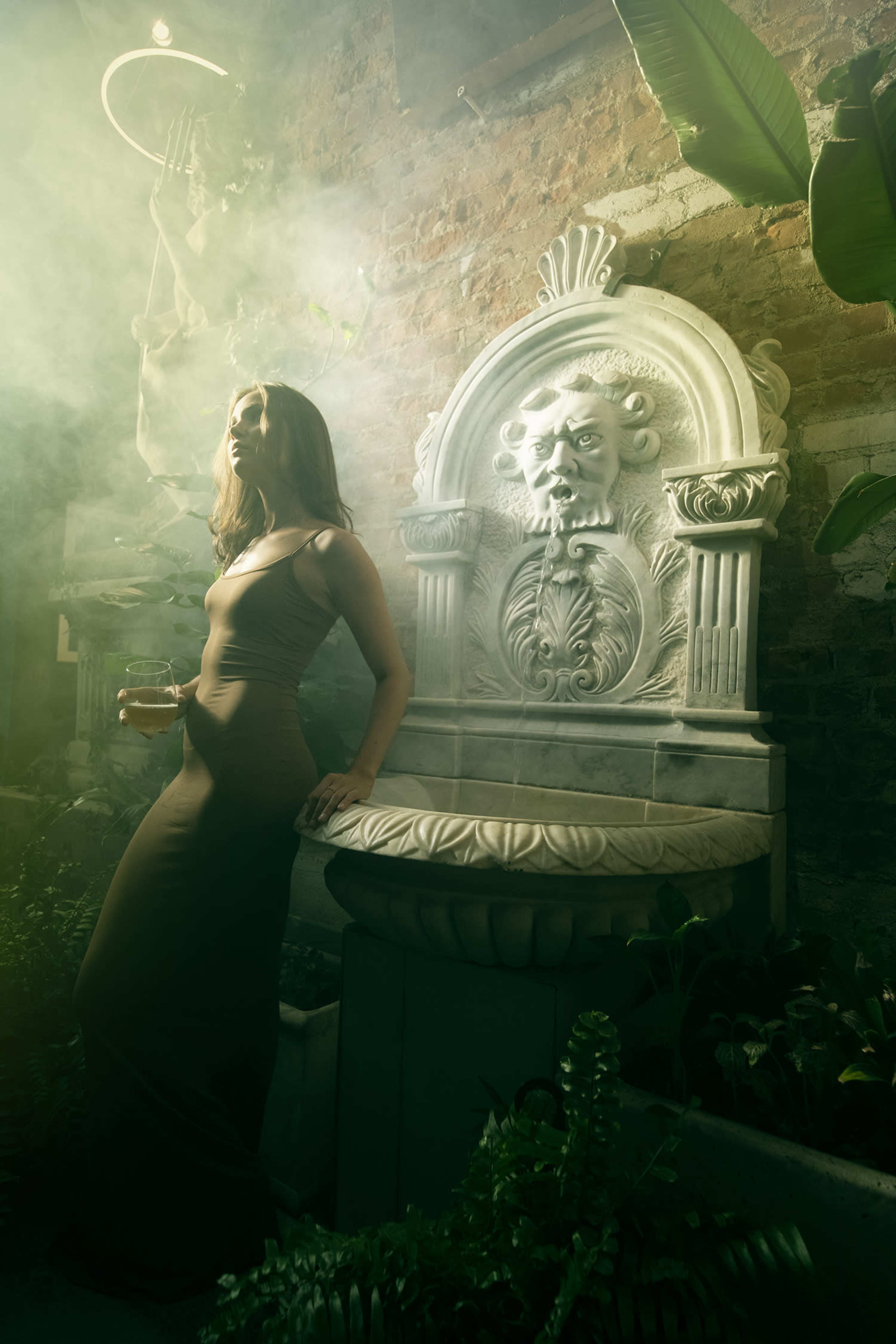 A woman in a fitted brown dress leans against a marble fountain adorned with a lion's head, surrounded by greenery and atmospheric mist.
