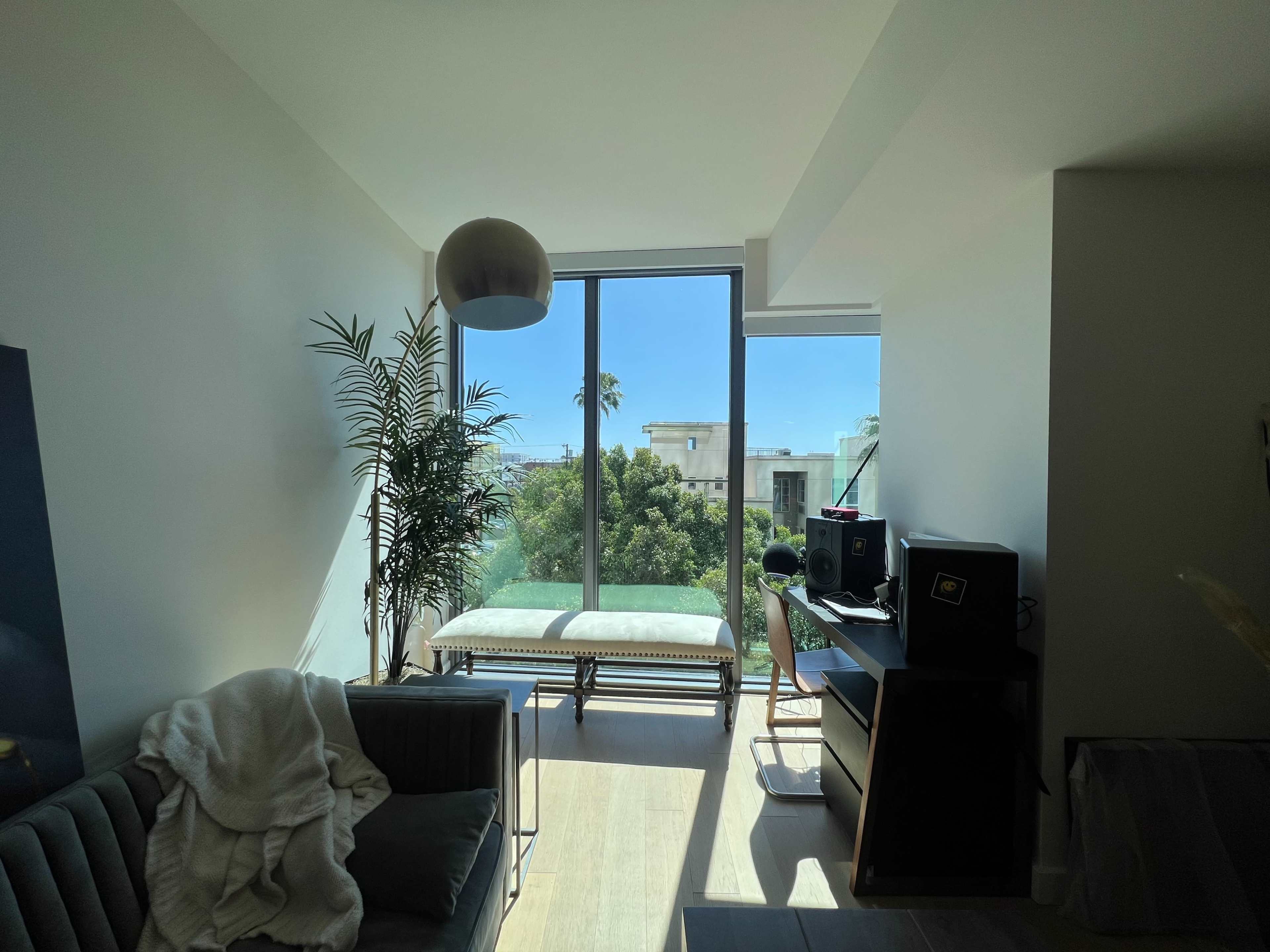 The image shows a bright living room with a large window, a couch, a desk with a computer setup, and greenery visible outside.