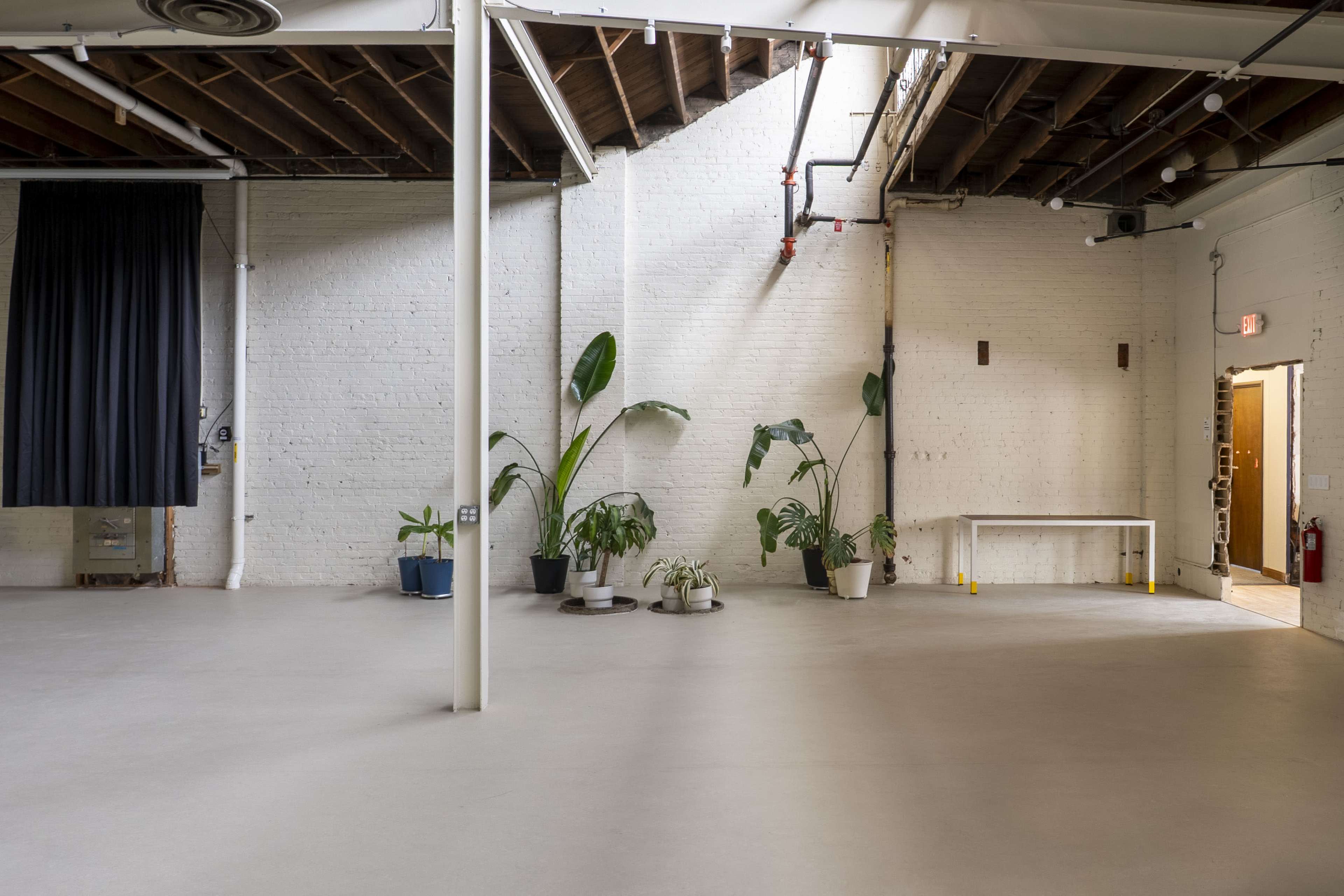The image shows a spacious, well-lit interior with white brick walls, polished concrete floors, and a few potted plants scattered around.