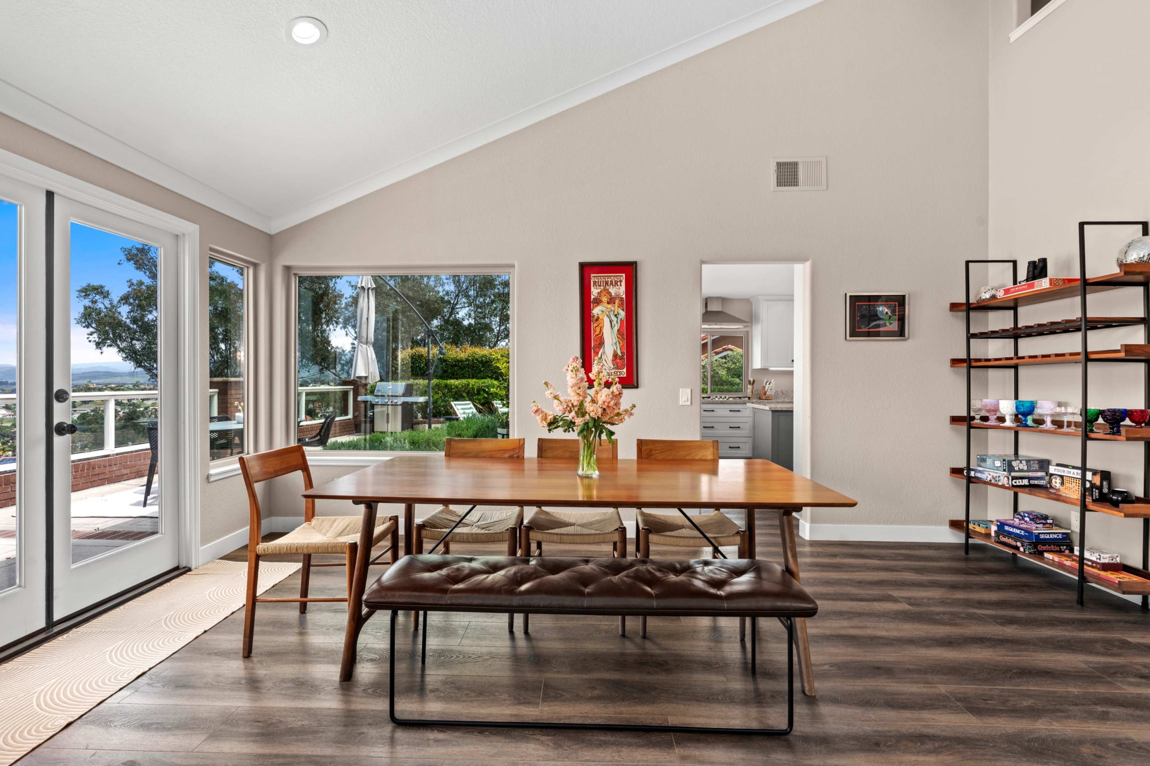 A dining area features a wooden table with chairs and a bench, surrounded by large windows showcasing an outdoor view.