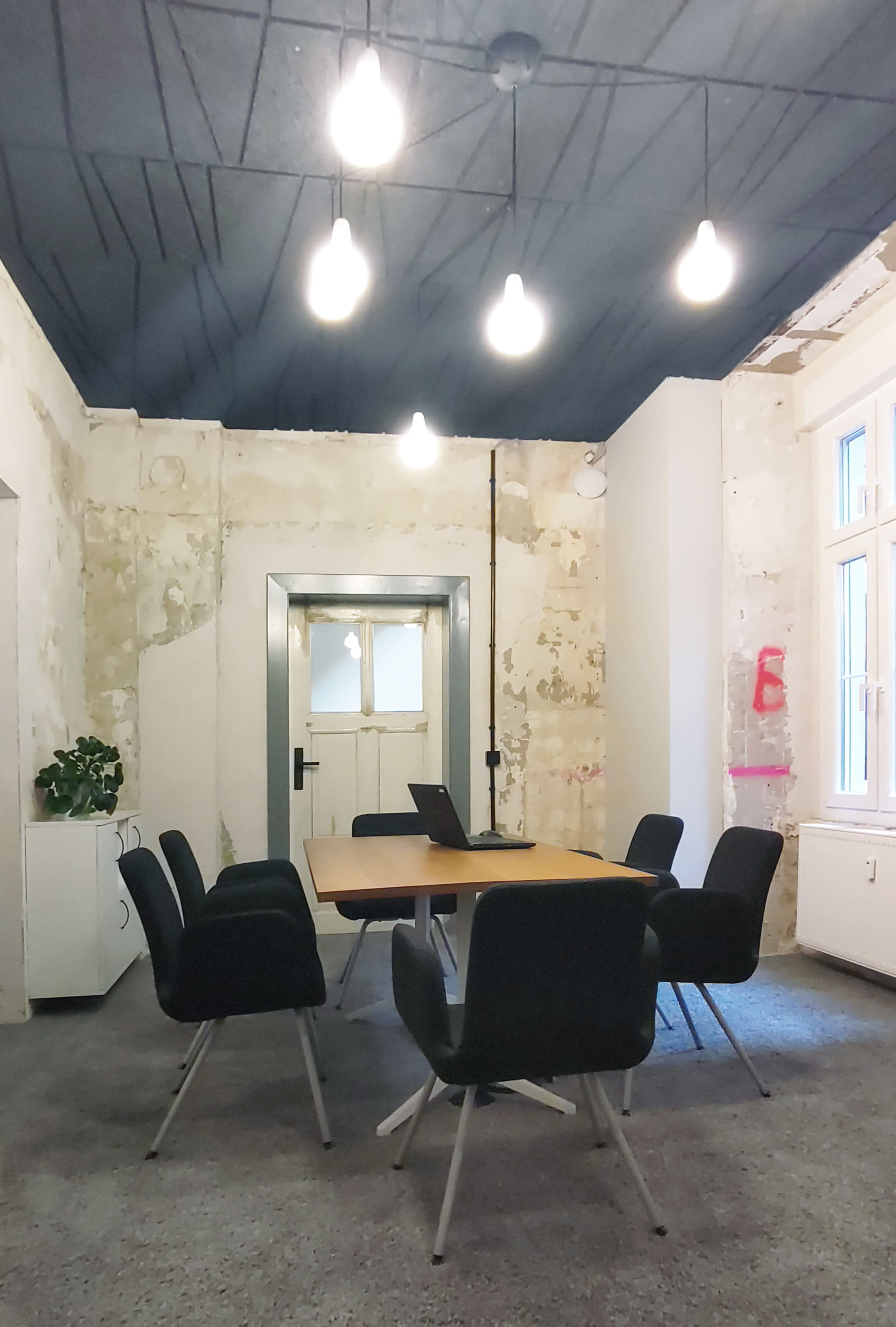 A conference room features a central wooden table surrounded by six chairs, with a laptop on the table and exposed light bulbs hanging from the ceiling.