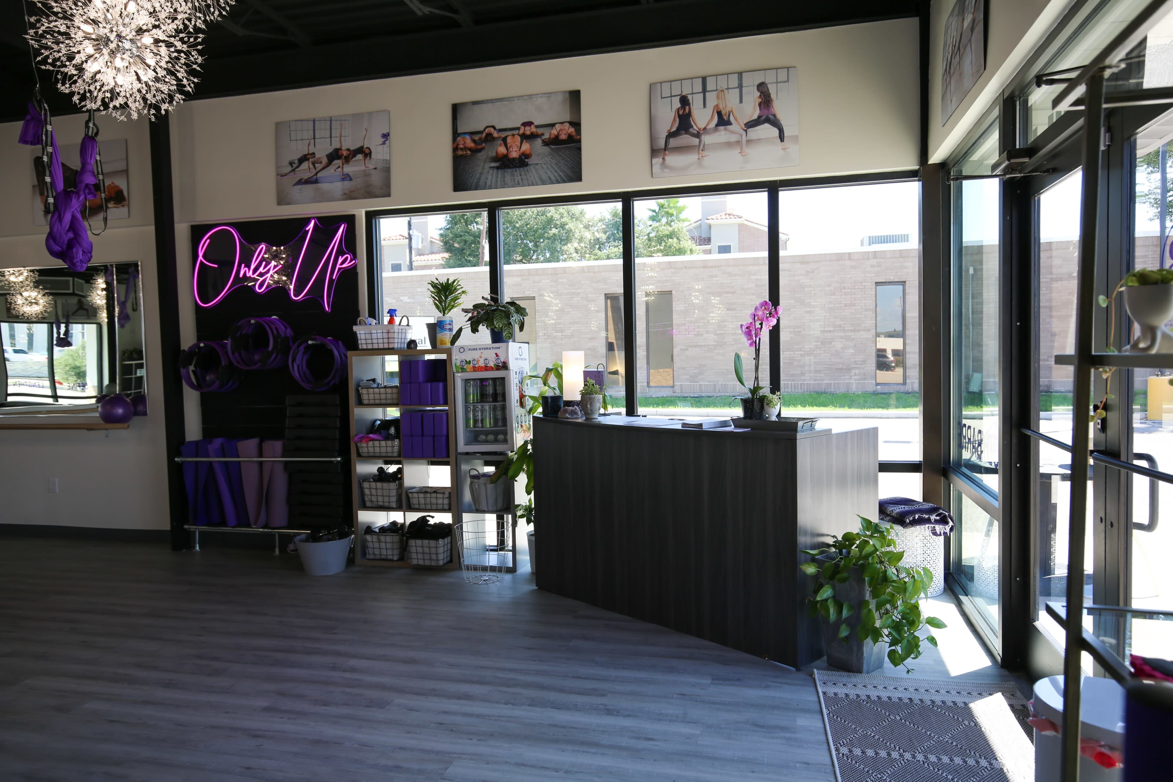 The image shows the interior of a fitness studio with large windows, a front desk, decorative plants, and purple-themed decor.
