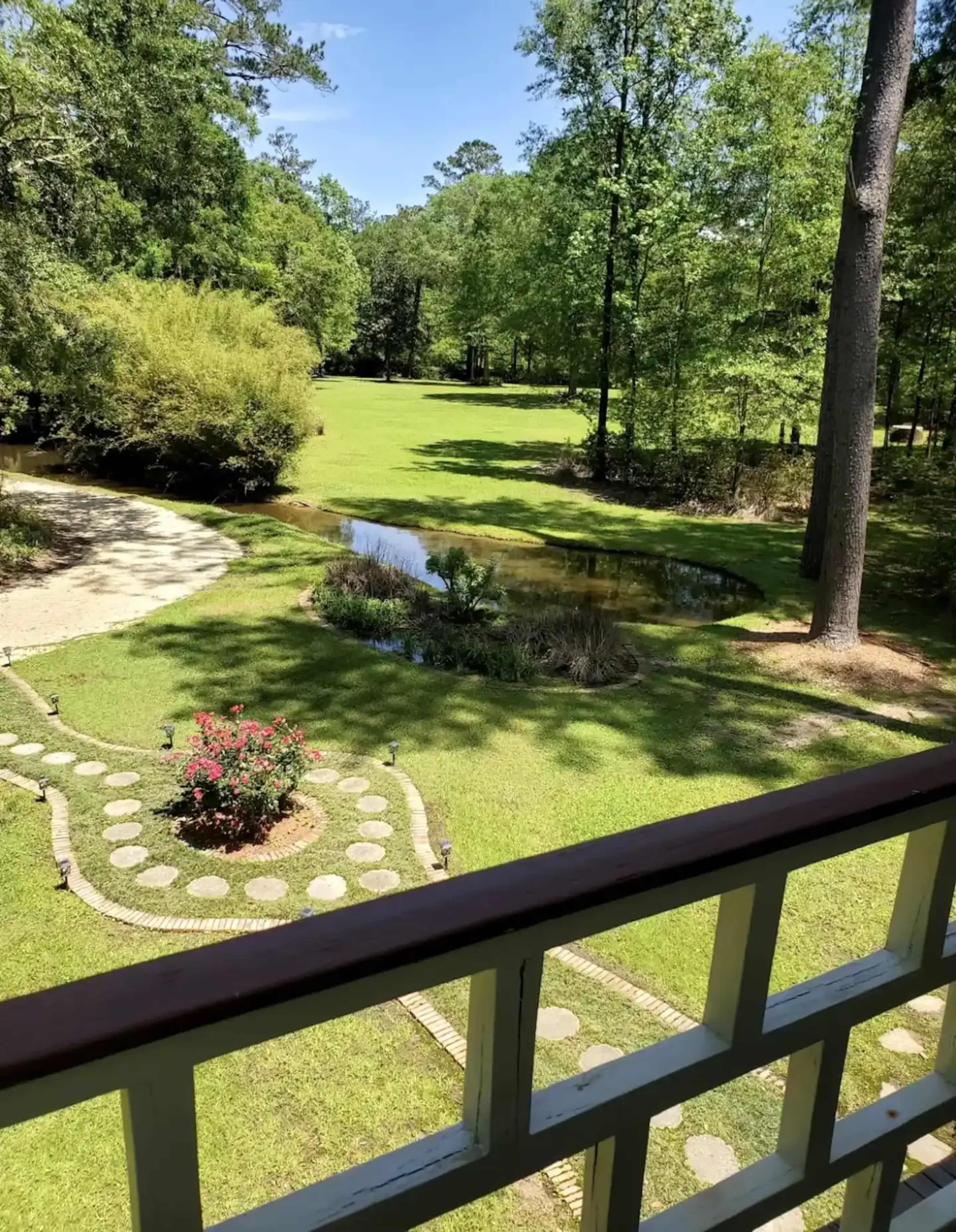 A green landscape featuring a manicured garden with a flower bed, a small pond, and trees in the background.