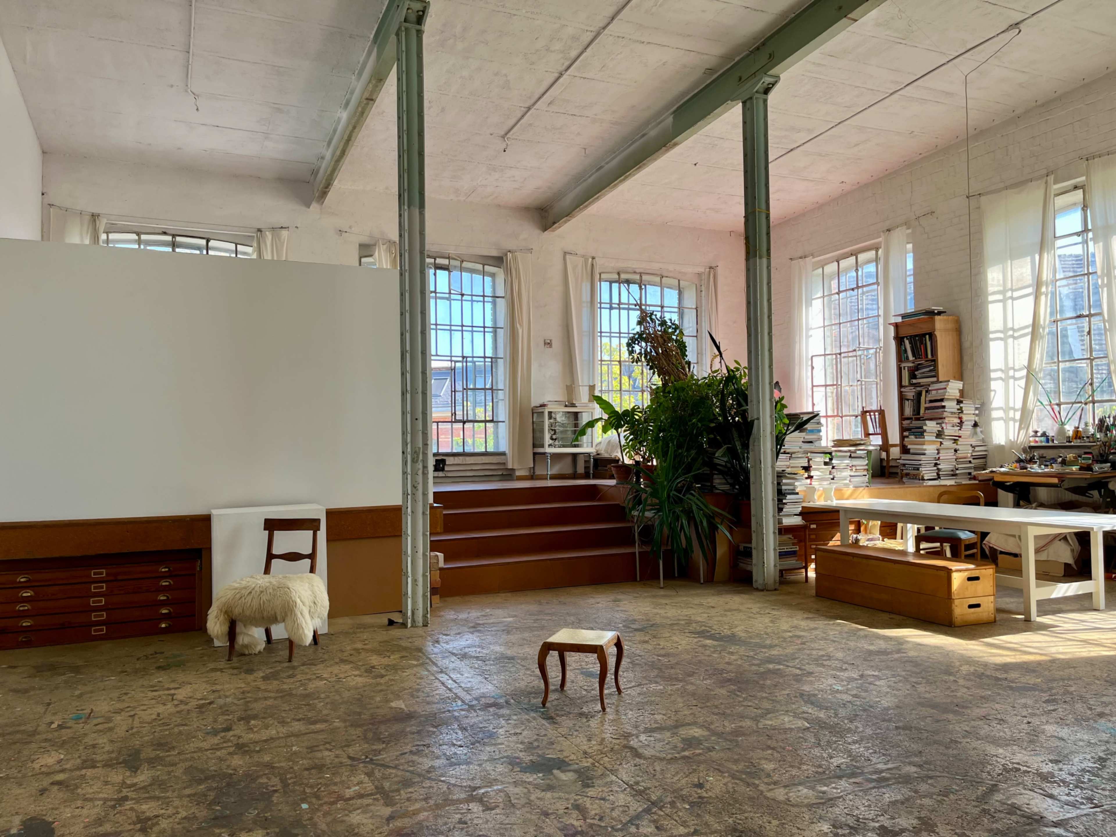 The image depicts a spacious studio with large windows, a bare wooden floor, and various furniture pieces, including a small table and shelving filled with books.
