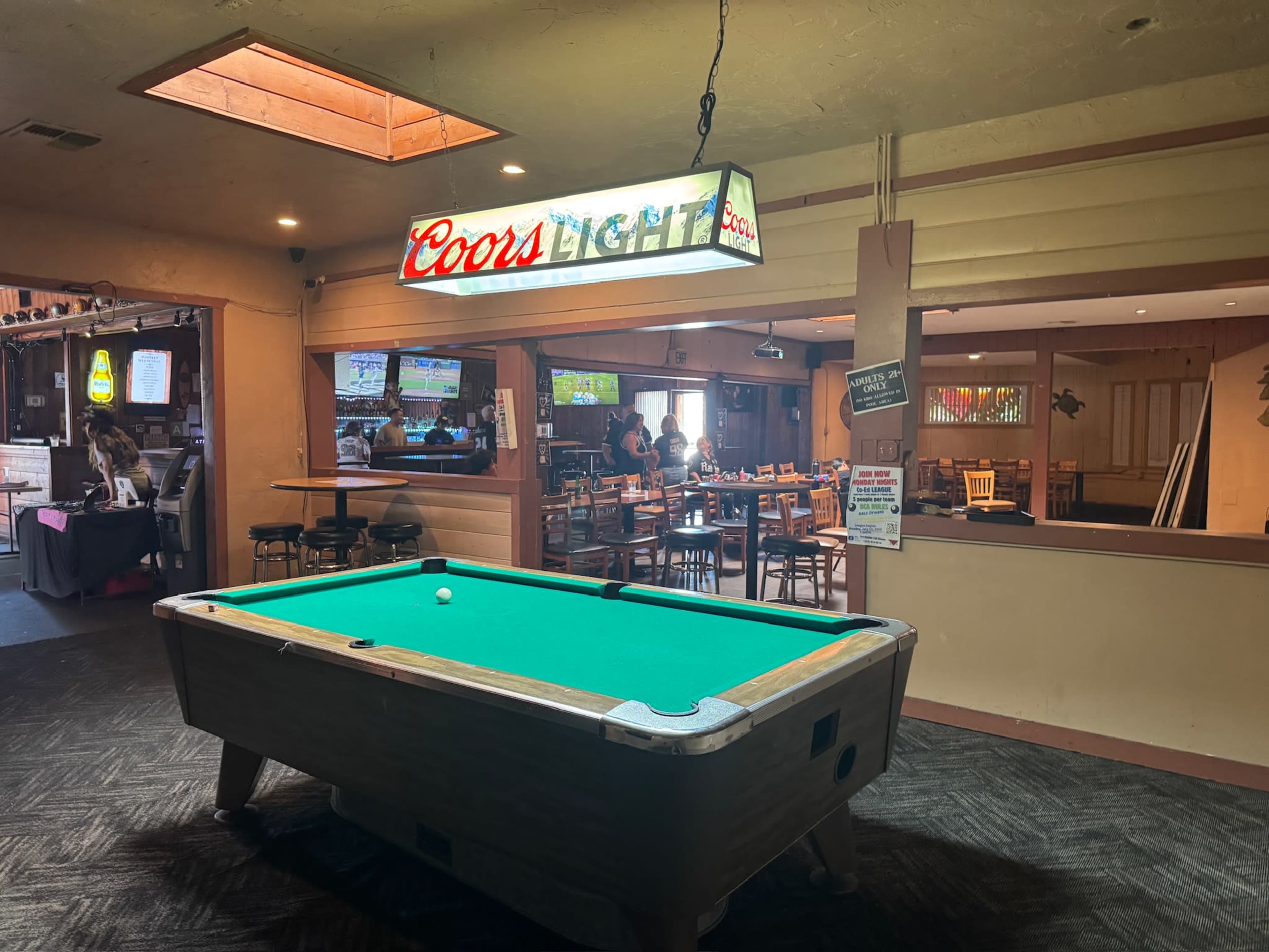 A pool table is centered in a dimly lit room featuring a Coors Light sign, with a dining area visible in the background.