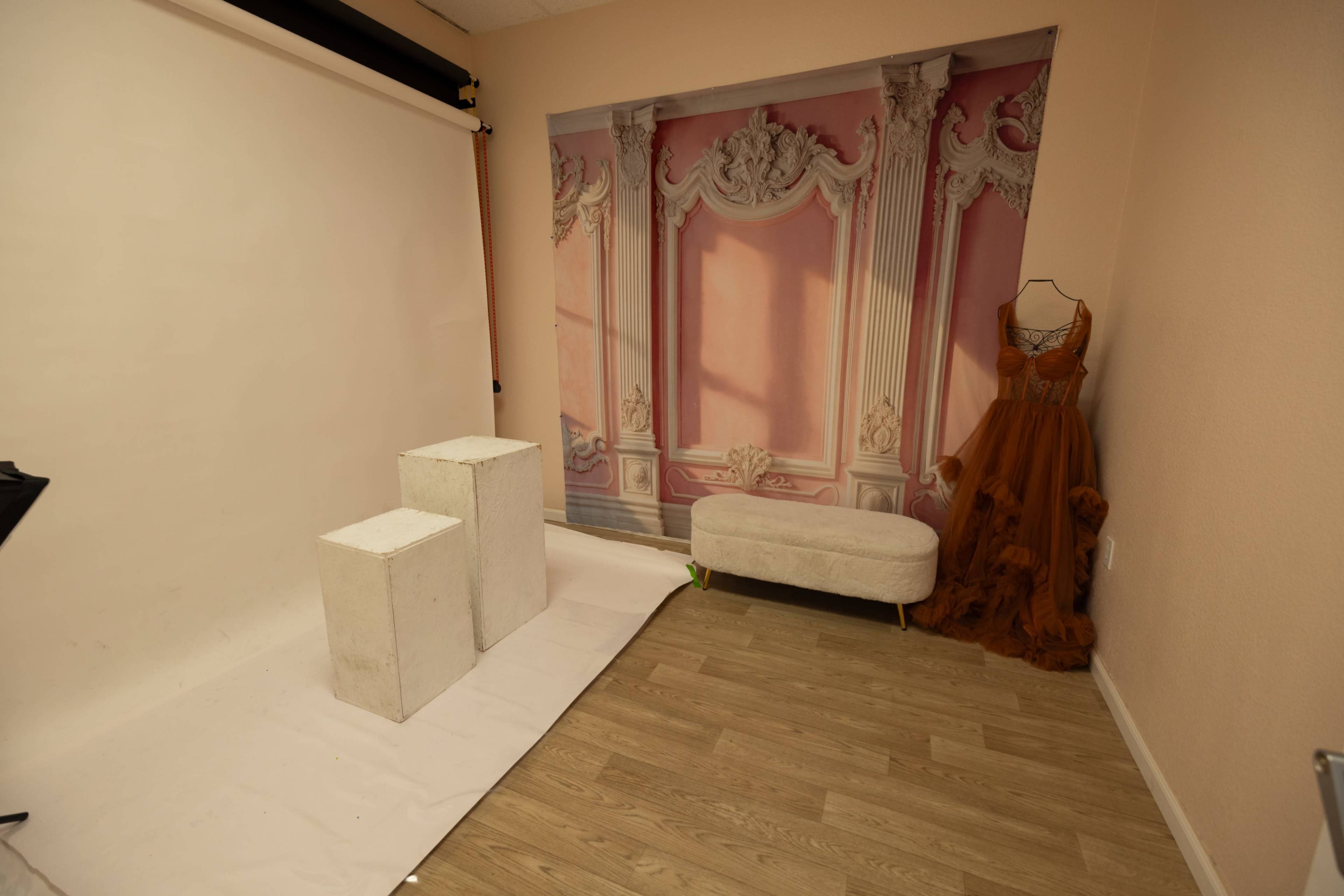 The image shows a photography studio featuring a beige backdrop with architectural details, two white pedestals, a light-colored bench, and an evening gown displayed on a mannequin.