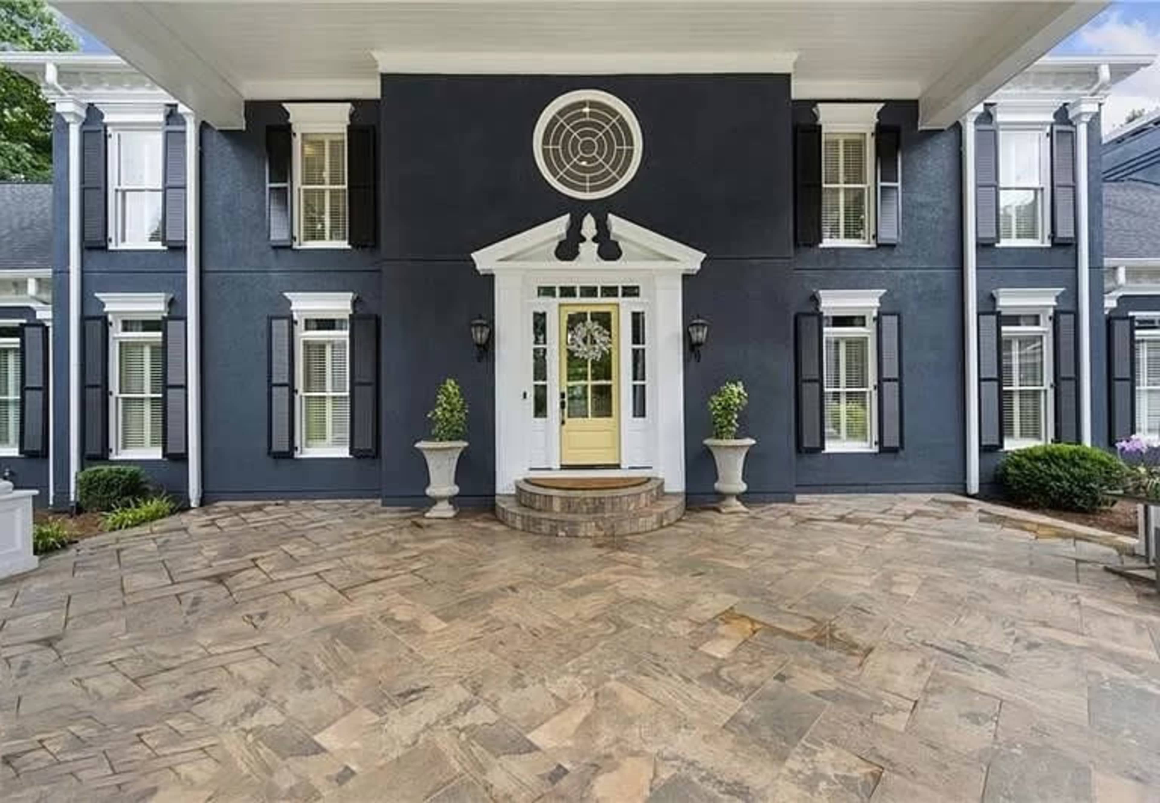 The image shows an elegant entrance of a two-story house with a circular design feature above the doorway, flanked by symmetrical columns and black shutters.