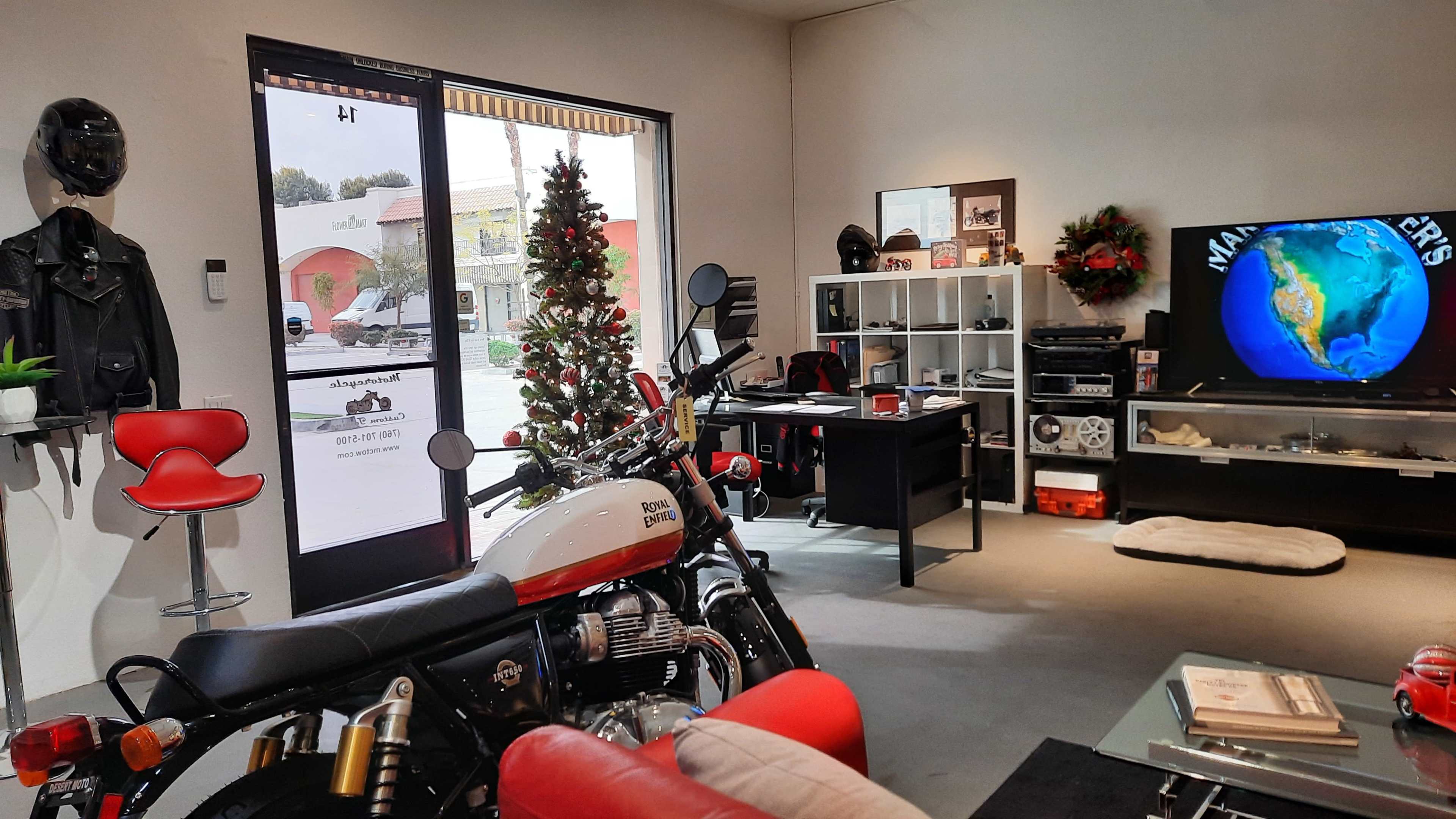 A modern motorcycle shop featuring a Royal Enfield motorcycle near a desk, a television displaying a globe, and a decorated Christmas tree.