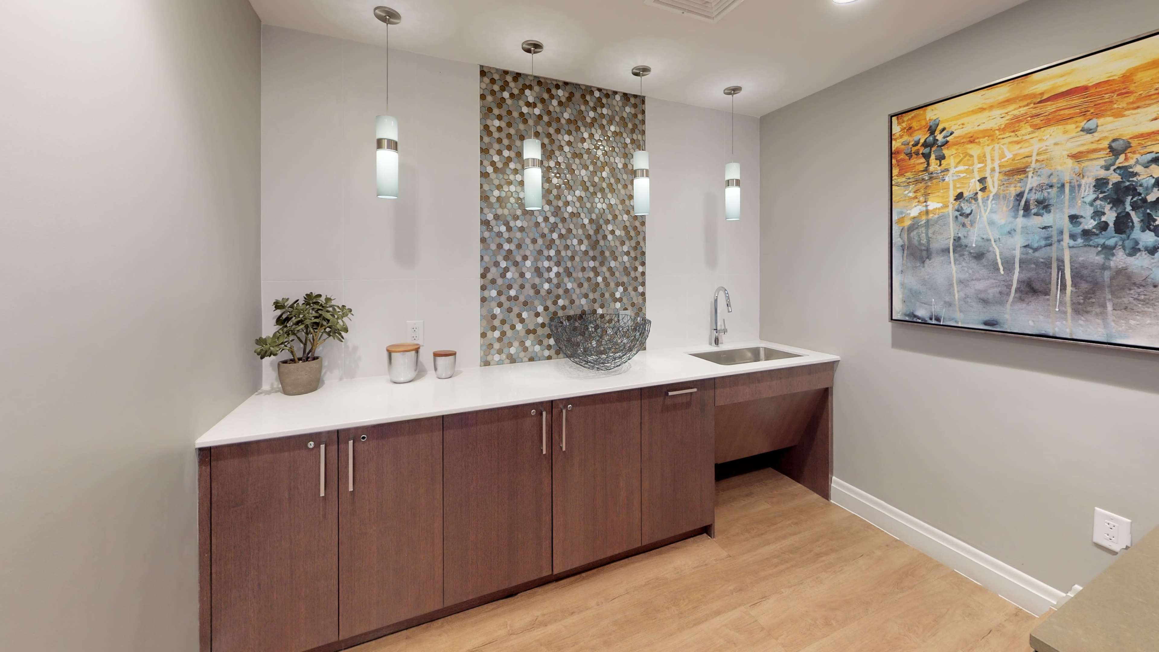 A modern kitchenette features a wooden cabinet below a minimalist countertop with a sink, decorative items, and a mosaic wall.