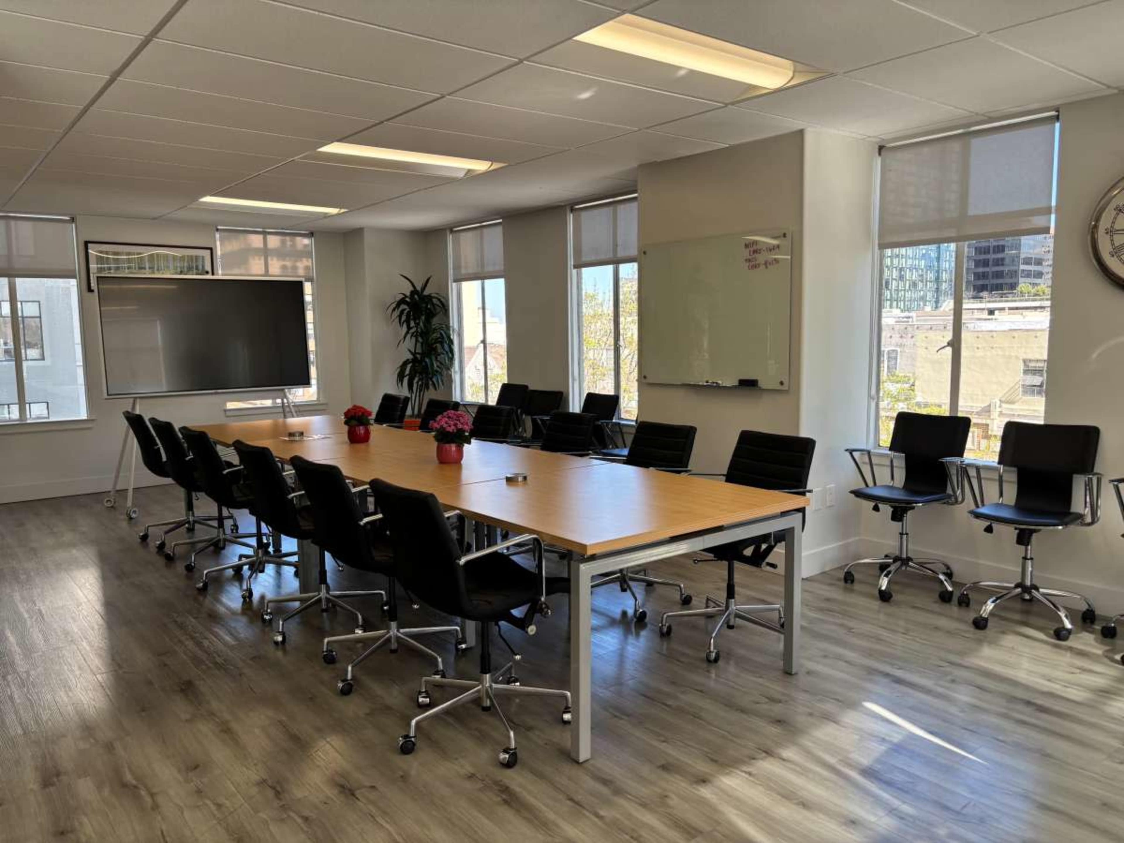 The image shows a modern conference room with a long wooden table, black chairs, a large screen, and windows providing natural light.