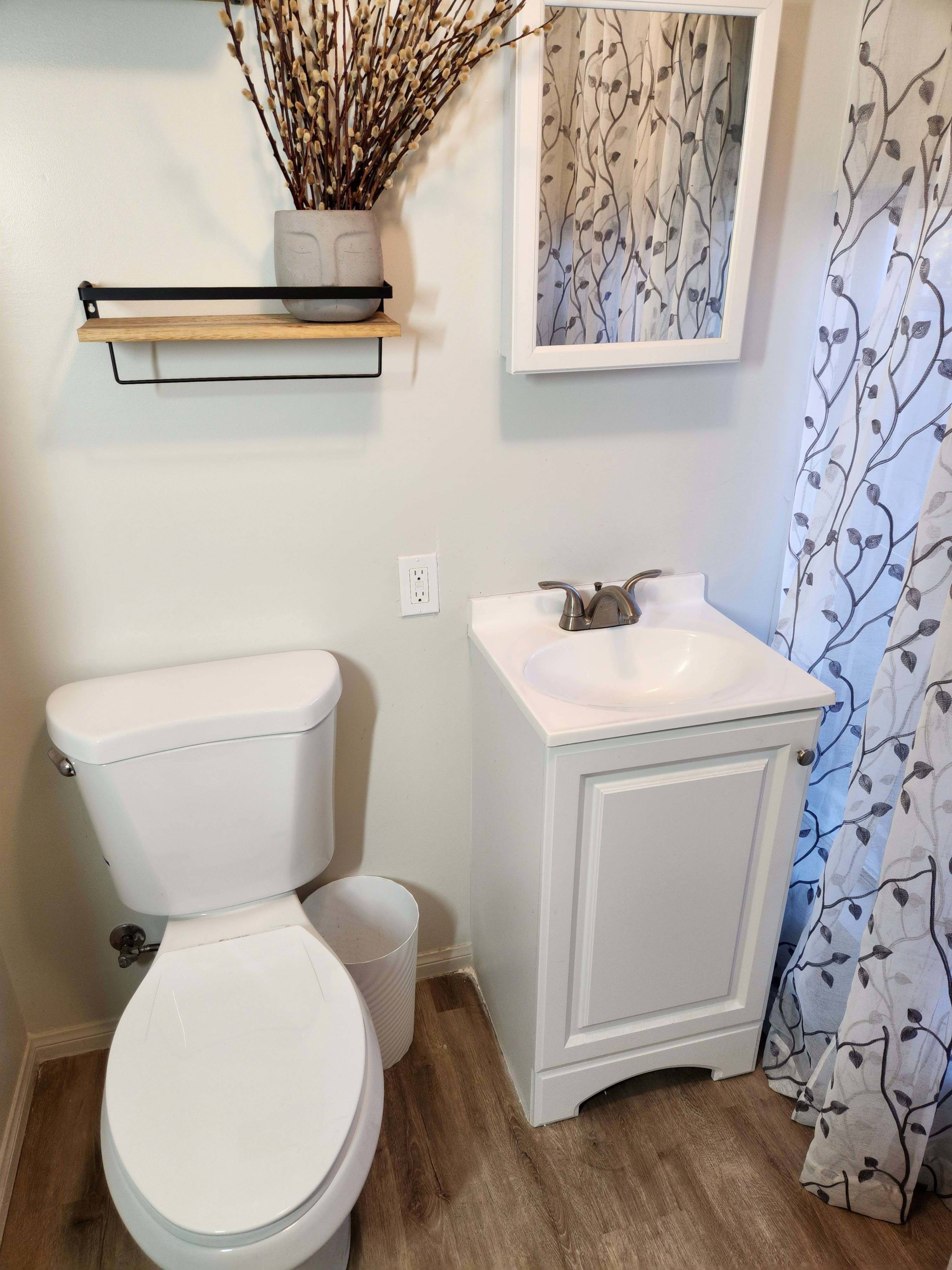 A small bathroom with a white toilet, a sink with a cabinet beneath it, a wall-mounted shelf holding a decorative vase, and sheer curtains with a floral pattern.