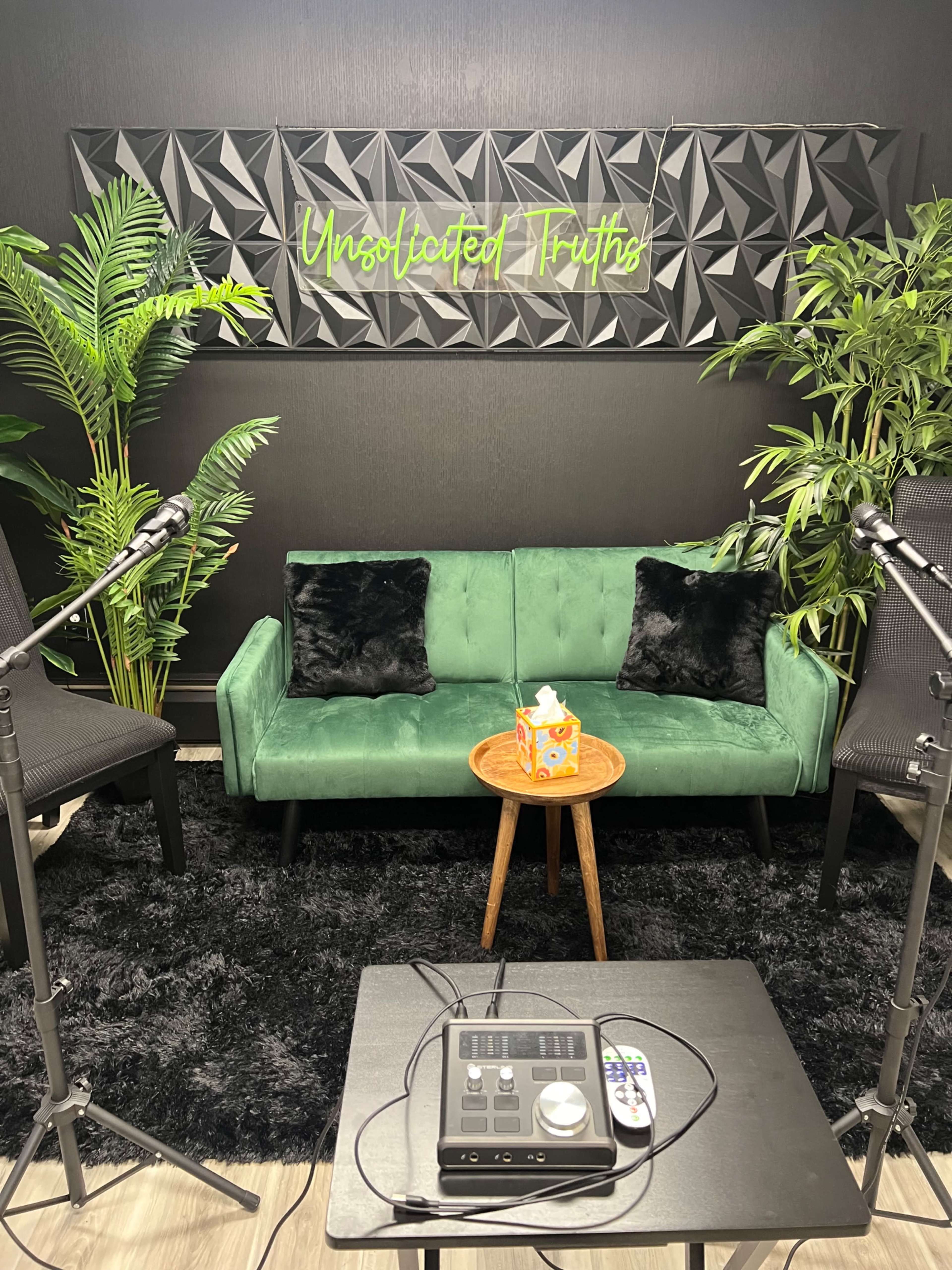 A modern podcast studio featuring a green couch with black pillows, a round wooden table, and plants, set against a black wall with the text "Unsolicited Truths" above.