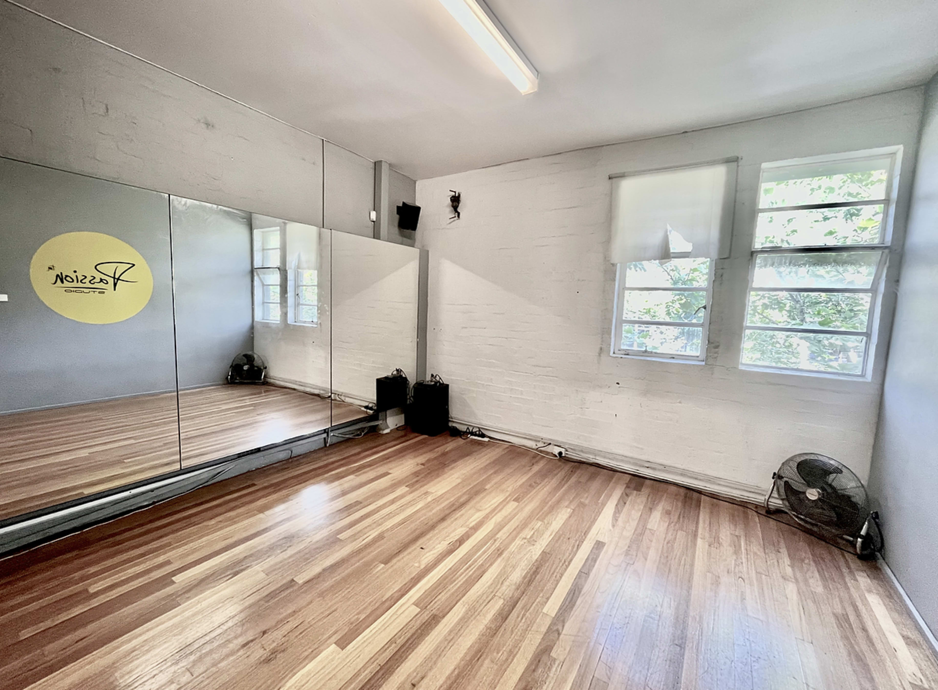 A mirrored wall reflects a bright dance studio with wooden flooring and large windows.