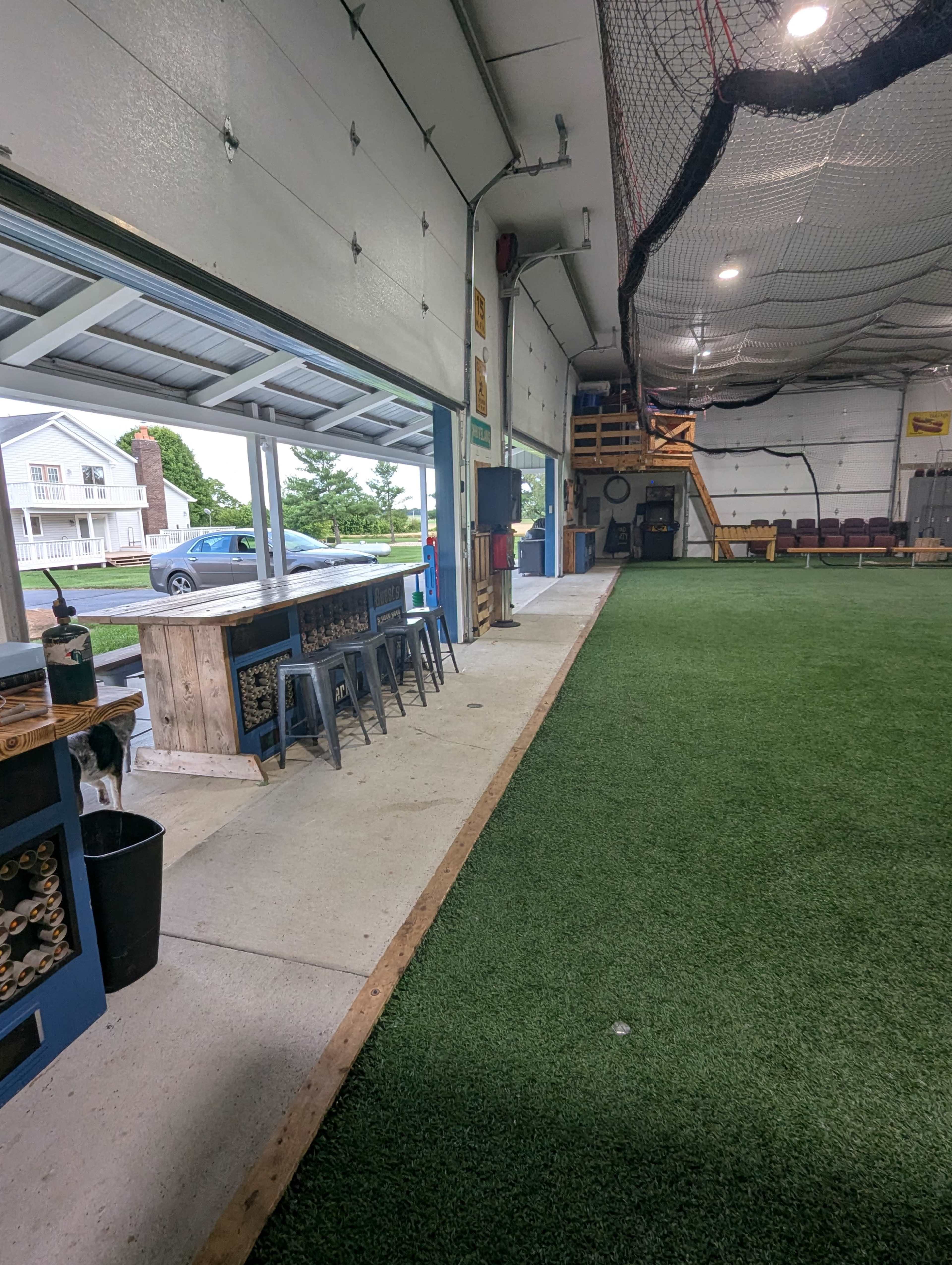 The image shows a spacious indoor training facility featuring artificial turf, a counter with stools, and a netted area for sports practice.