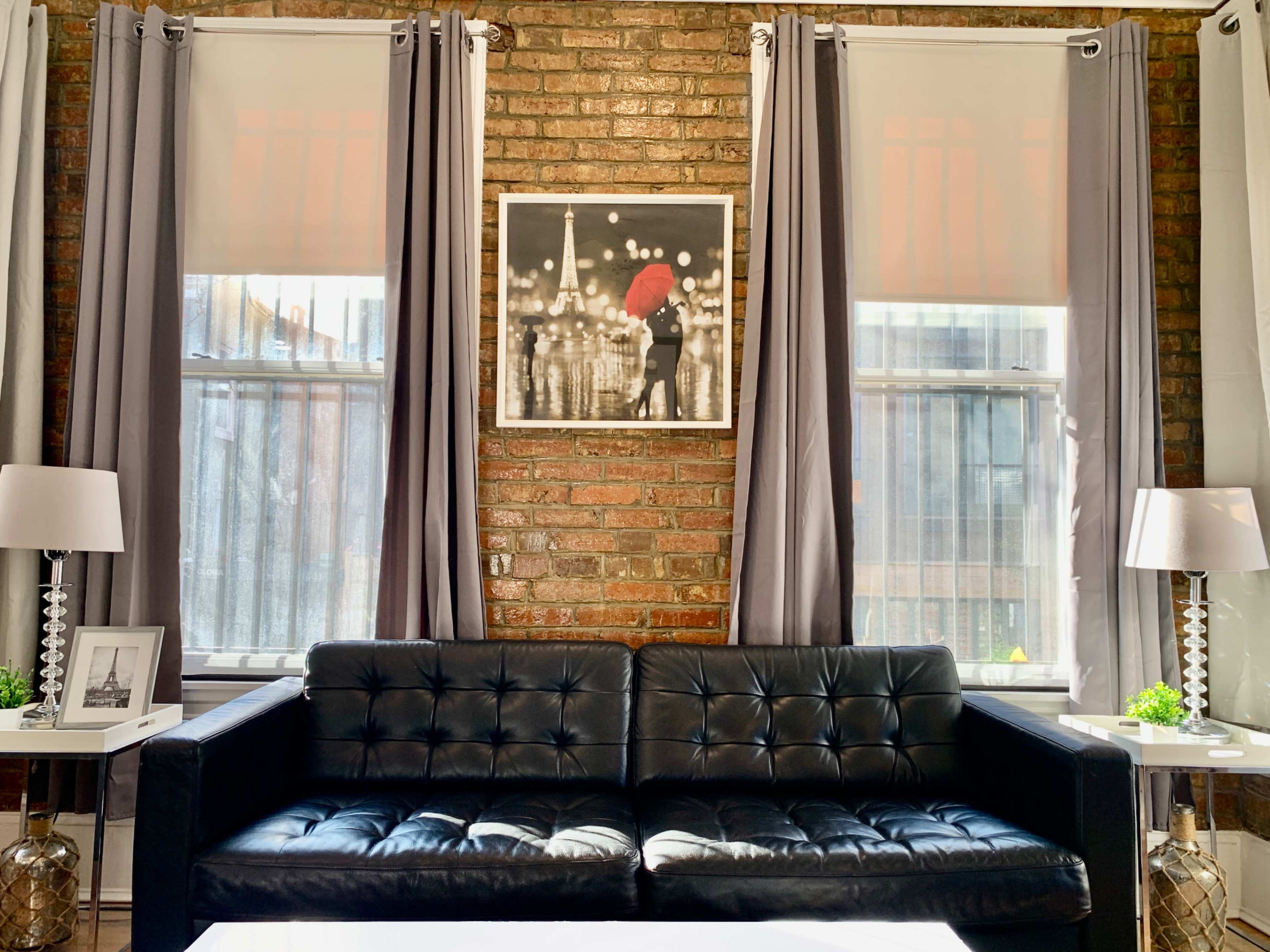 A living room with a black leather couch facing a framed photograph of a couple under an umbrella, flanked by tall windows with gray curtains.