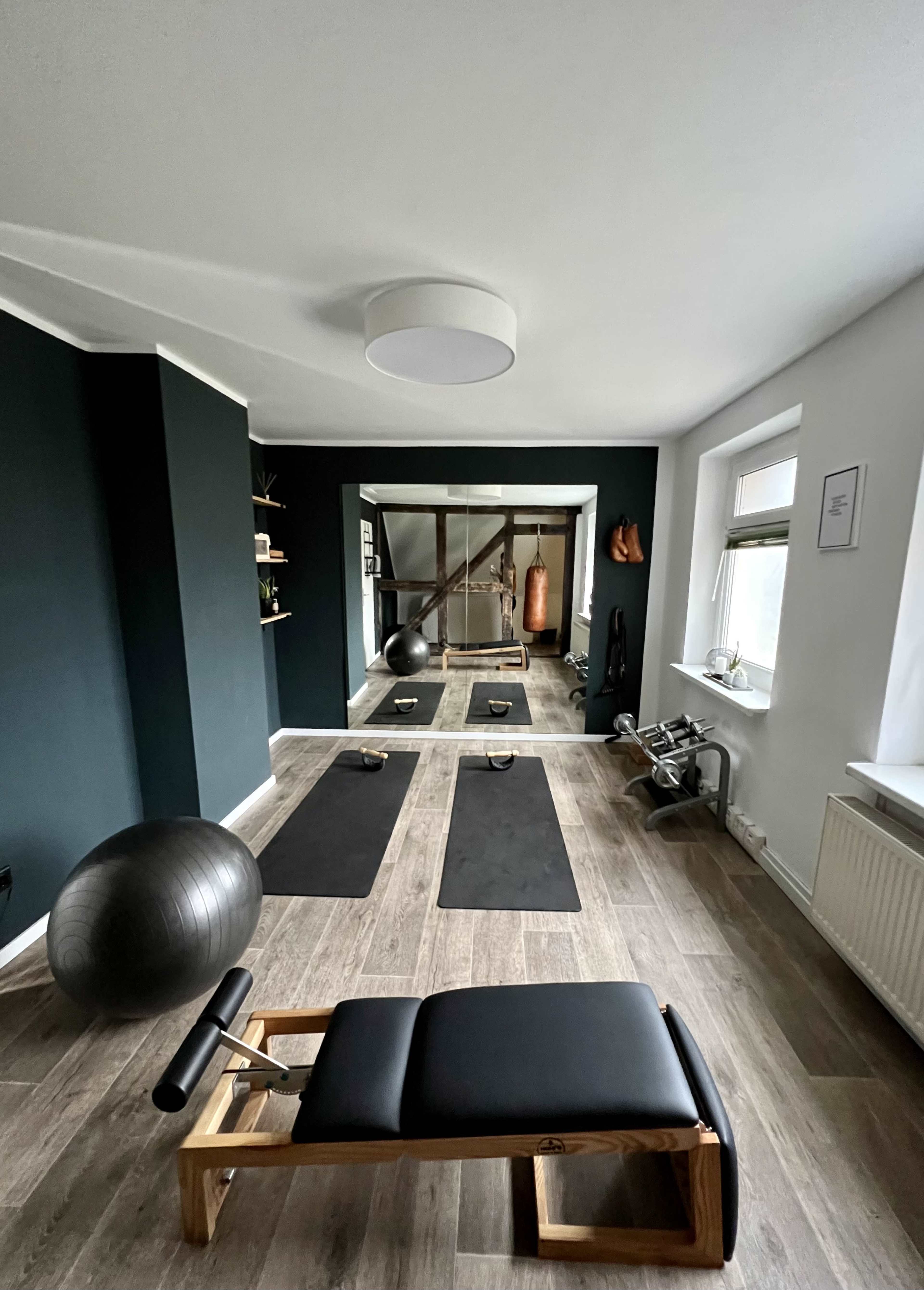 The image shows a home gym featuring a workout bench, exercise mats, a stability ball, and a mirrored wall with a punching bag and fitness equipment.