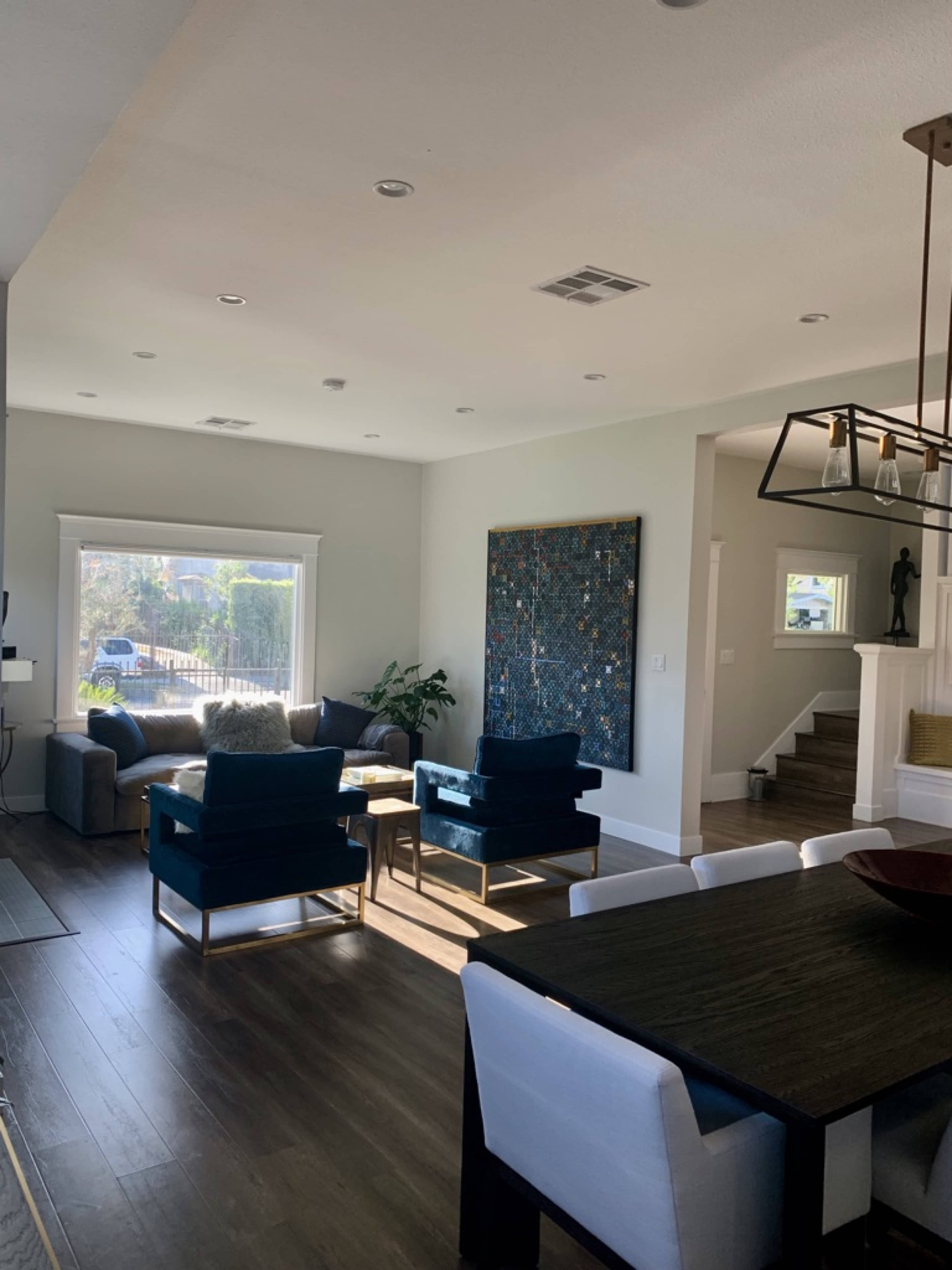 The image shows a modern living room and dining area featuring a combination of dark furniture, wood flooring, and a large wall art piece.