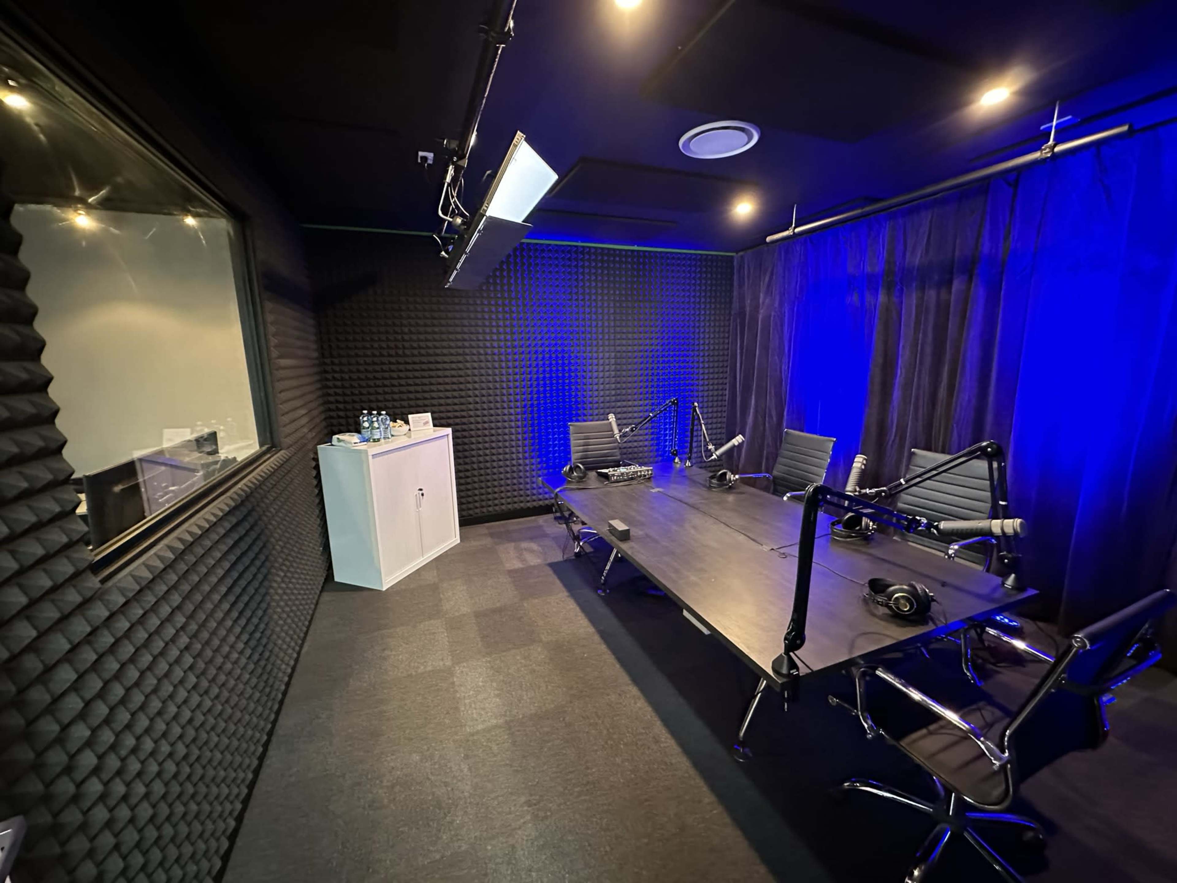 The image shows a soundproof recording studio featuring a large table with several chairs, microphones, and blue lighting.