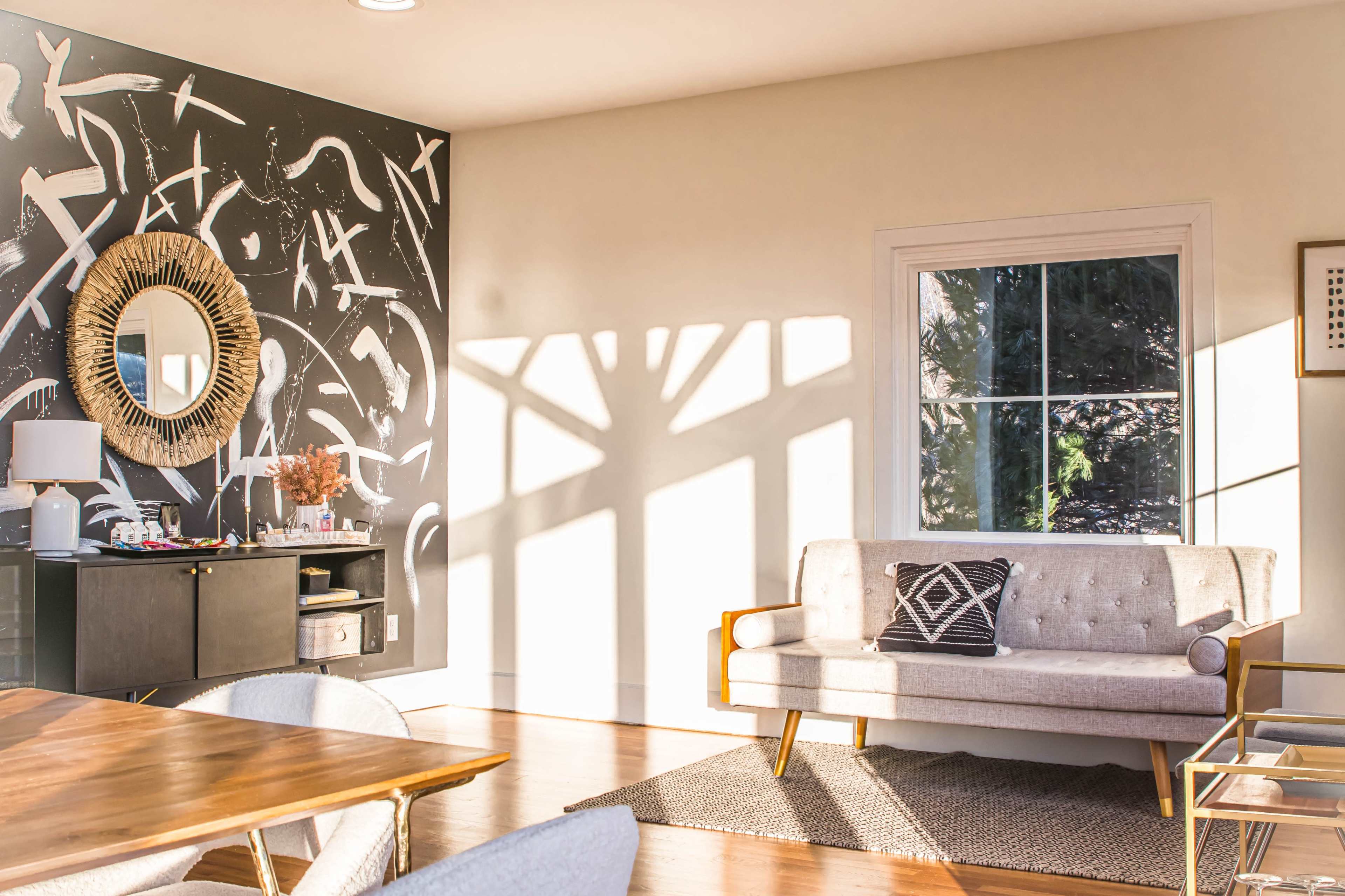 A living room with a geometric black and white wall art, a circular mirror, a gray sofa, and a sunlight-cast shadow through a window.