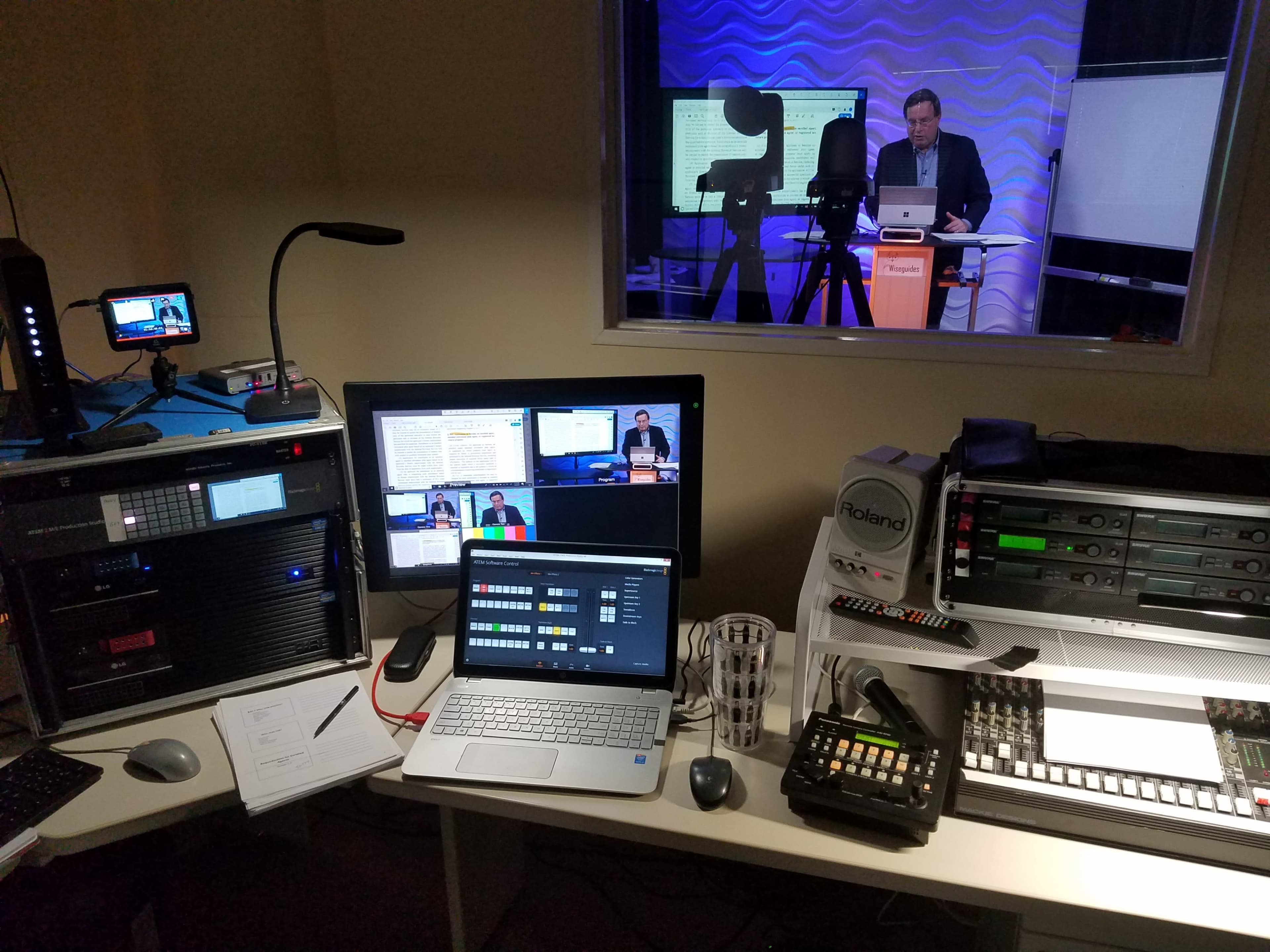 A multimedia control room with multiple screens displaying live video feeds and a presenter giving a presentation in a glass-walled studio.