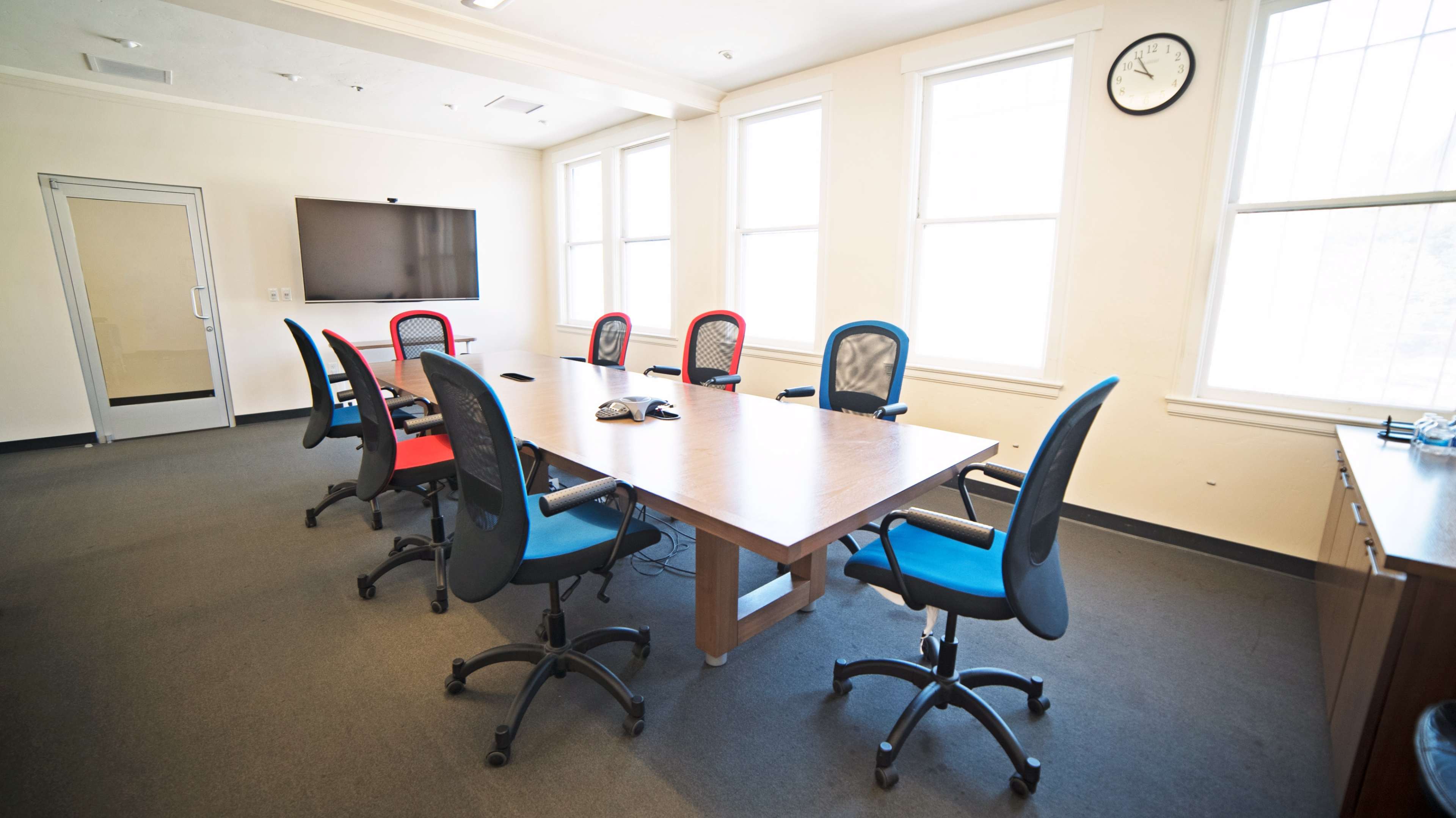 A conference room features a large wooden table surrounded by six office chairs in red and blue, with a television mounted on the wall and windows allowing natural light.