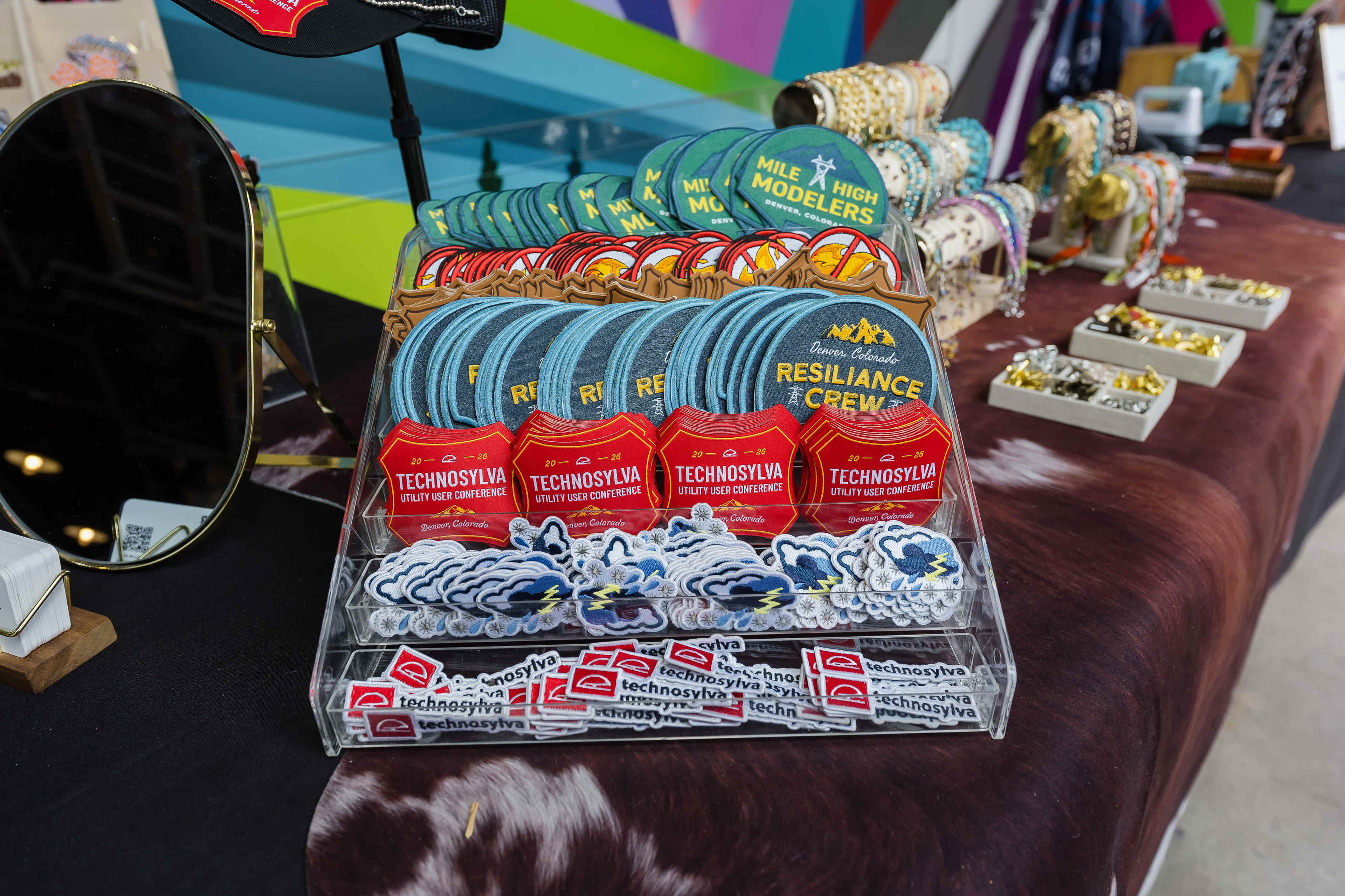 The image shows a display of colorful patches, including ones labeled "Resilience Crew" and "Technosylva," arranged neatly on a table at a market or event setting.