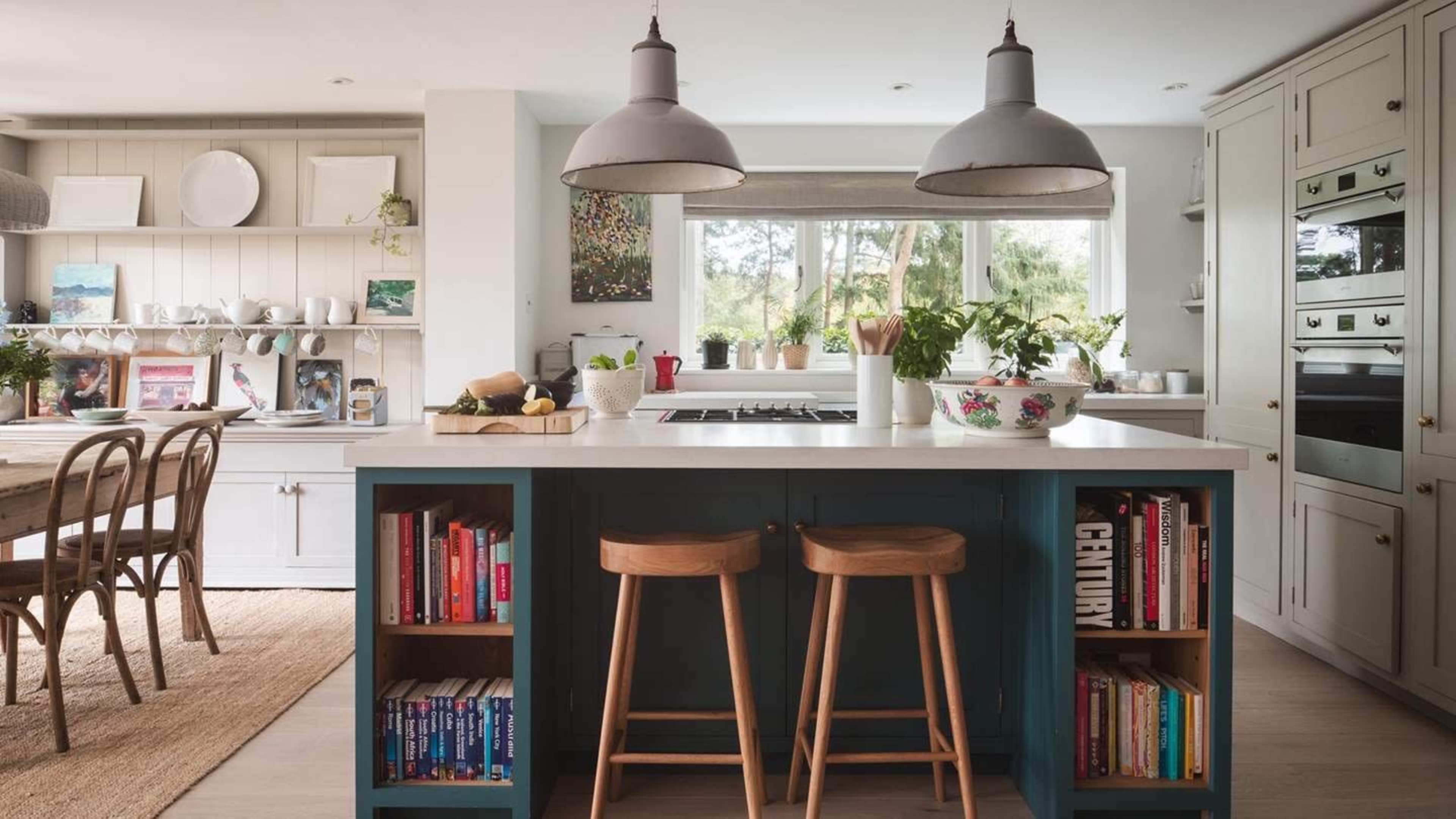 A modern kitchen features a central island with a stovetop, two wooden stools, and bookshelves underneath, while a dining area is visible in the background.