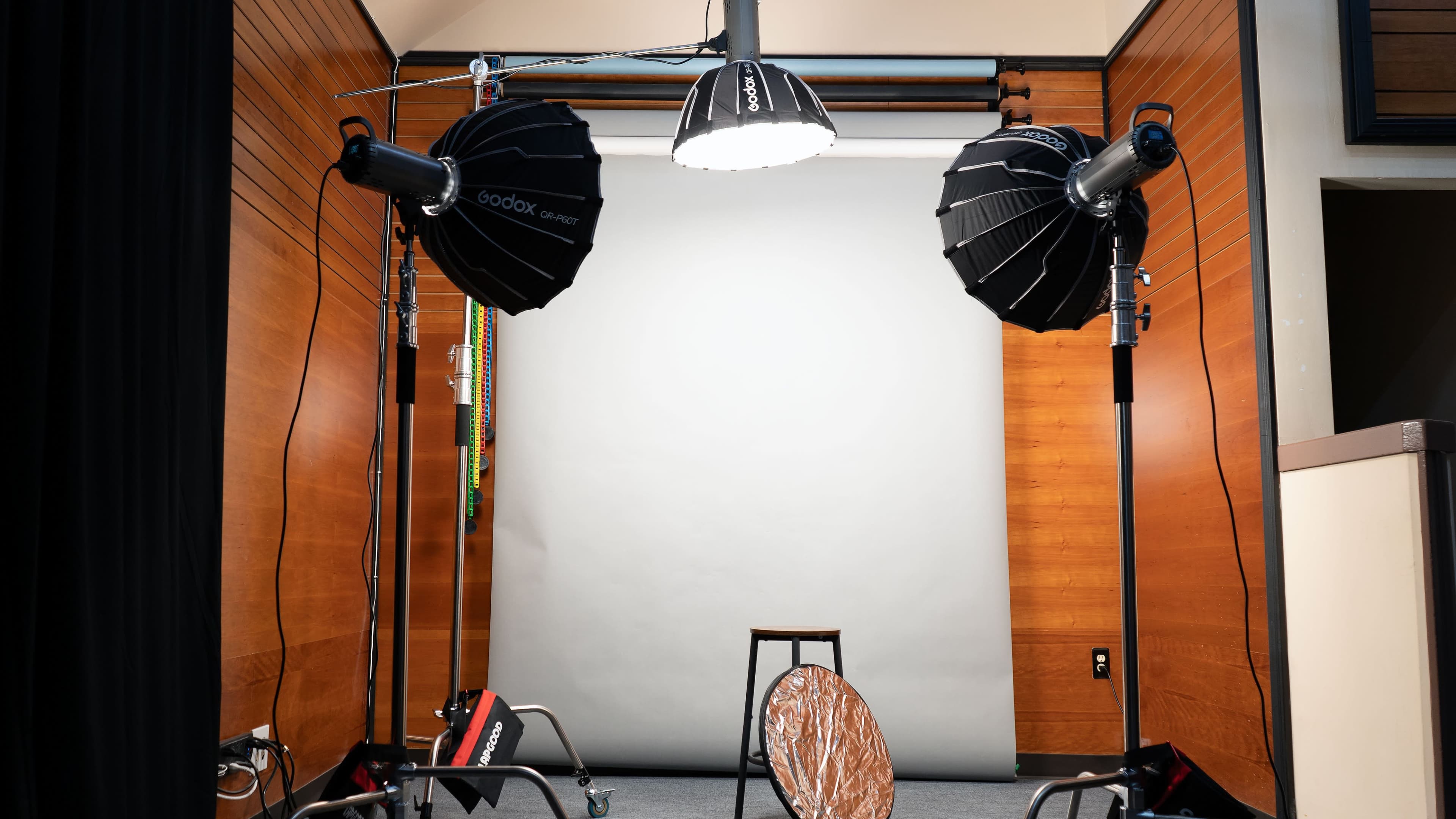 A photography studio setup with two softbox lights positioned in front of a blank backdrop, a stool in the center, and a reflective disc on the floor.