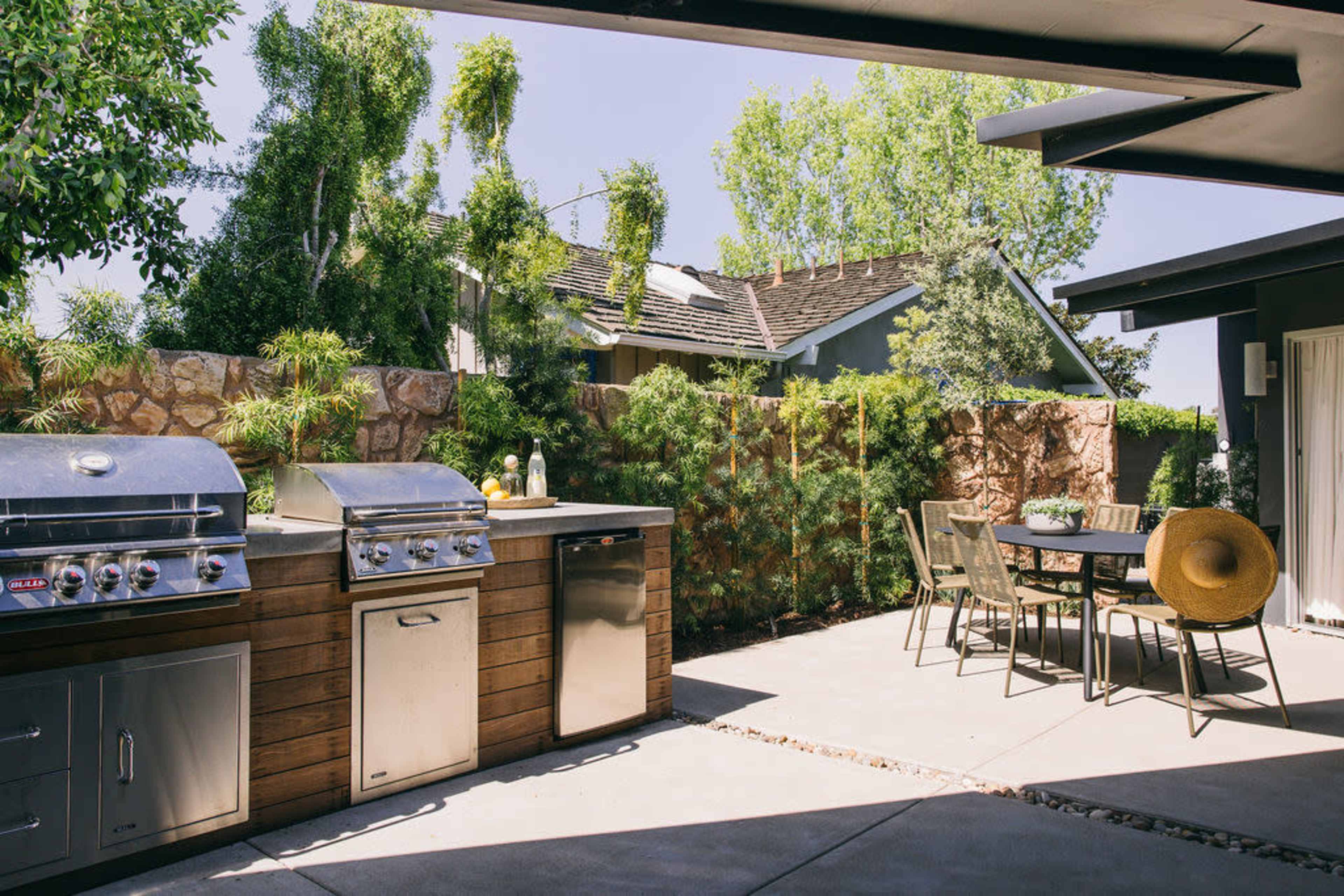 An outdoor kitchen area with two stainless steel grills and a dining table surrounded by greenery.