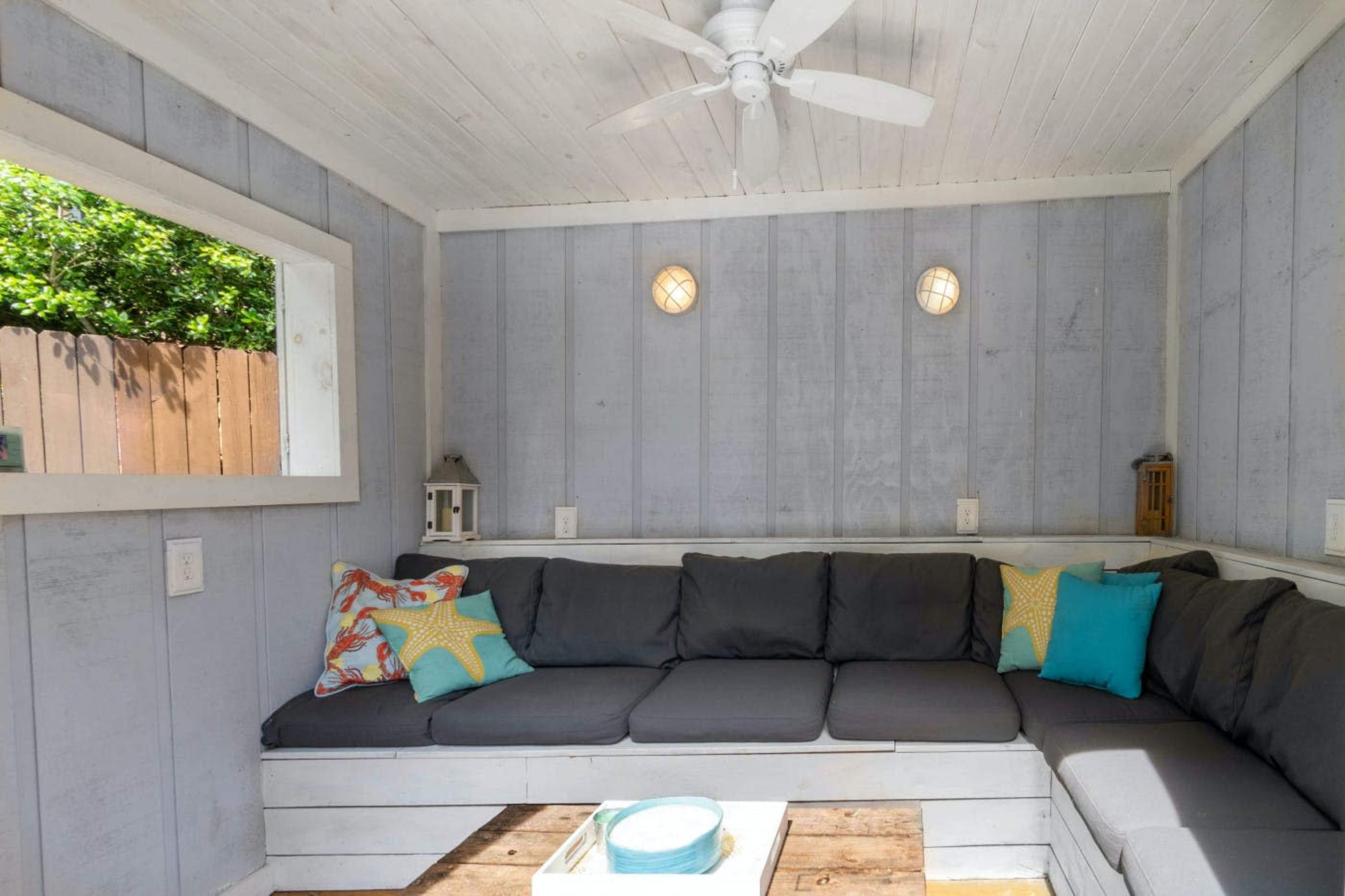 A cozy seating area with a gray sectional couch, colorful pillows, a central table, and a window allowing natural light.