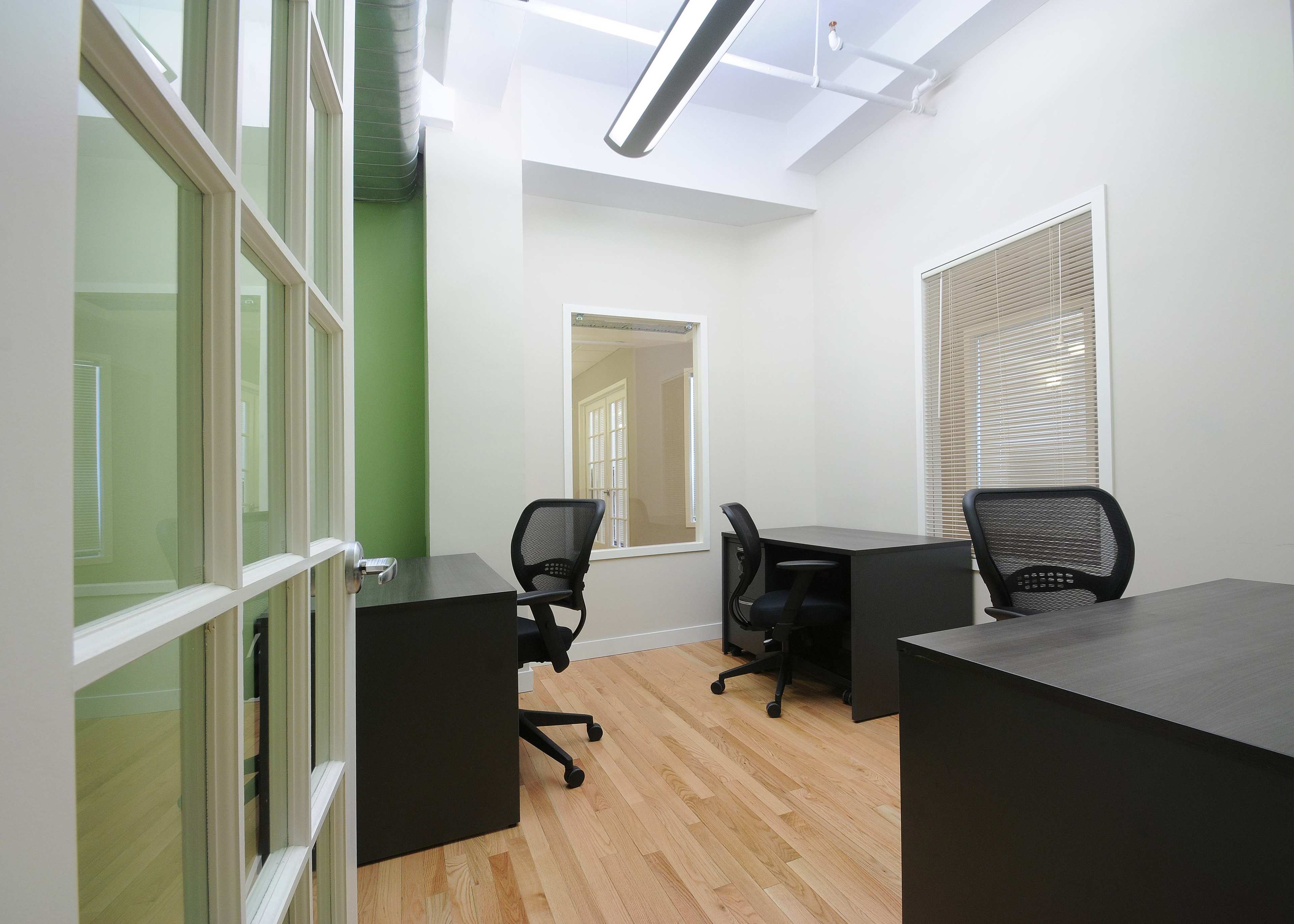 A modern office space with two black desks and rolling chairs, featuring light hardwood floors and green accent walls.