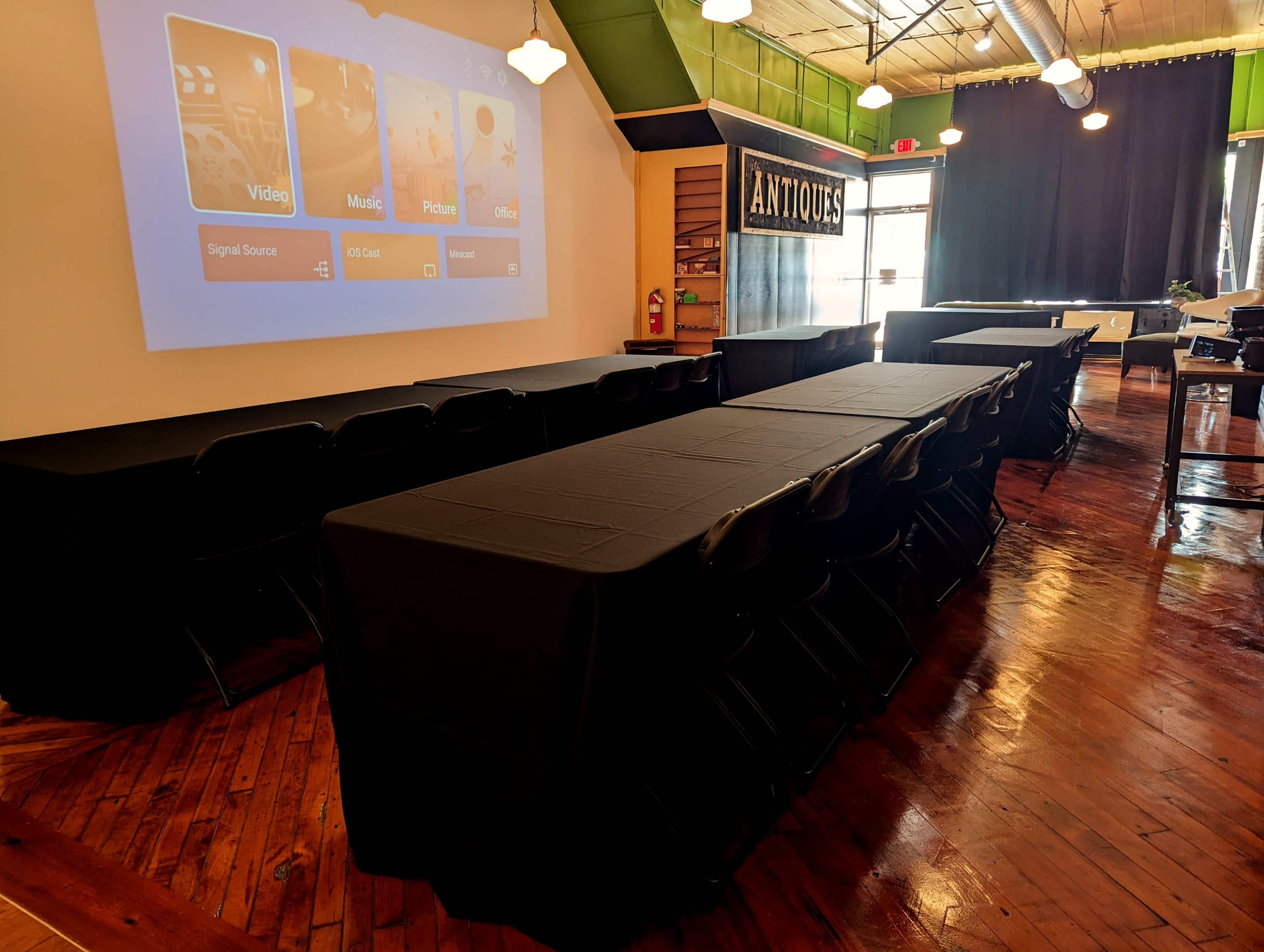 A room set up for a meeting, featuring several black rectangular tables facing a projection screen displaying options for video, music, pictures, and office.