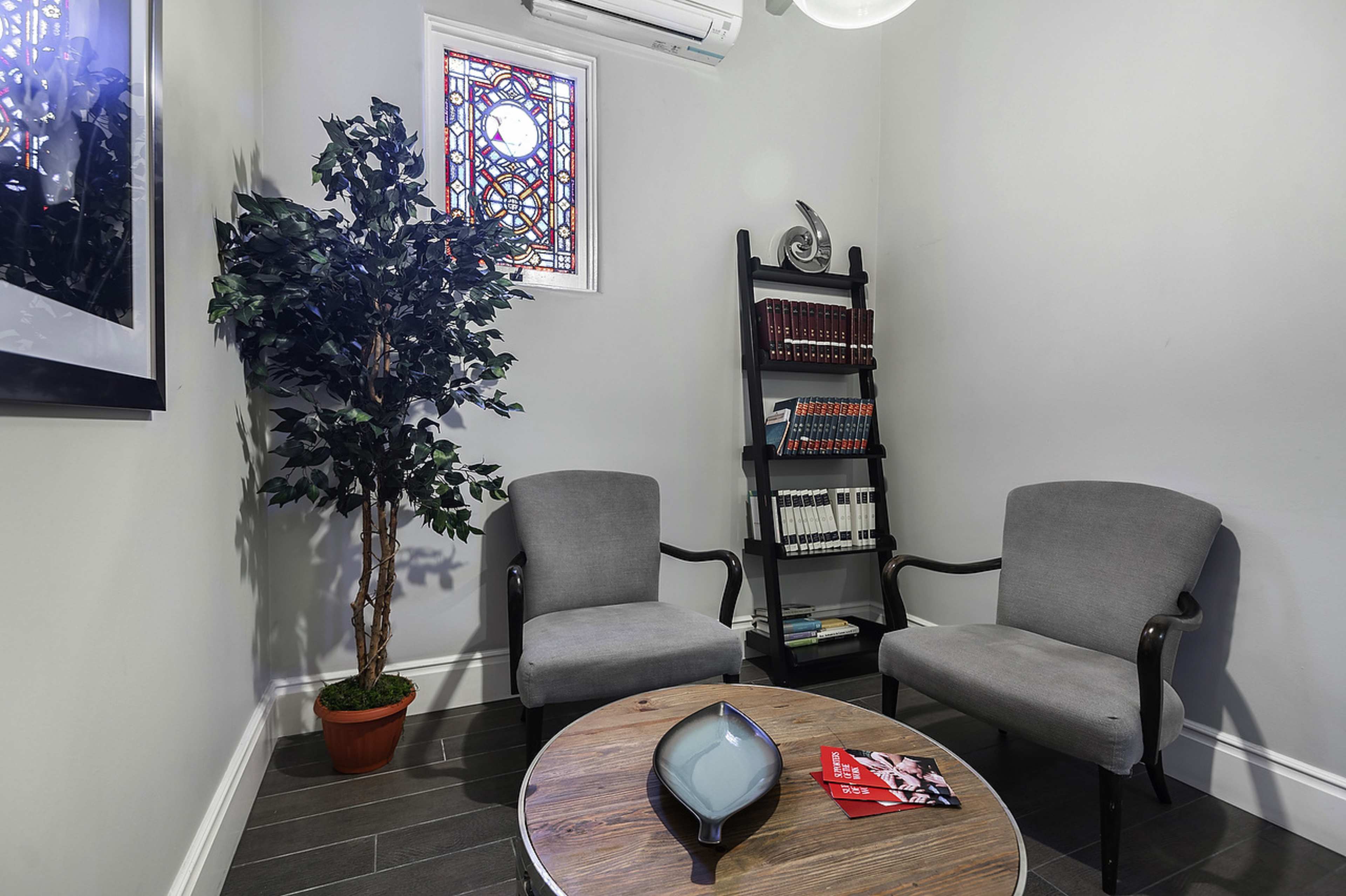 A small sitting area with two gray armchairs, a round wooden table, a bookshelf filled with books, and a decorative potted plant near a stained glass window.