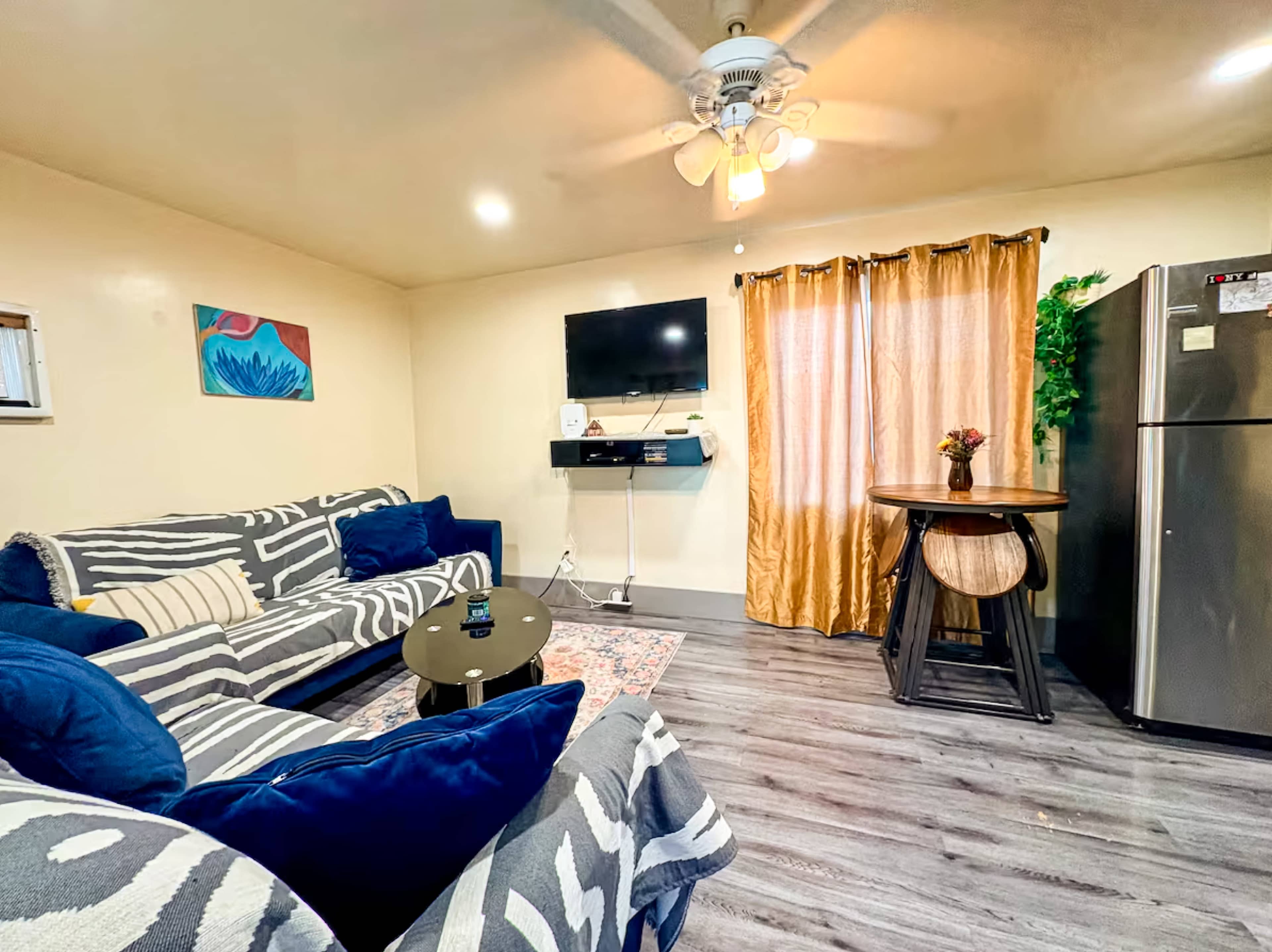 The image depicts a living room with two sofas, a mounted TV, a table, and a refrigerator, featuring a window draped with golden curtains.