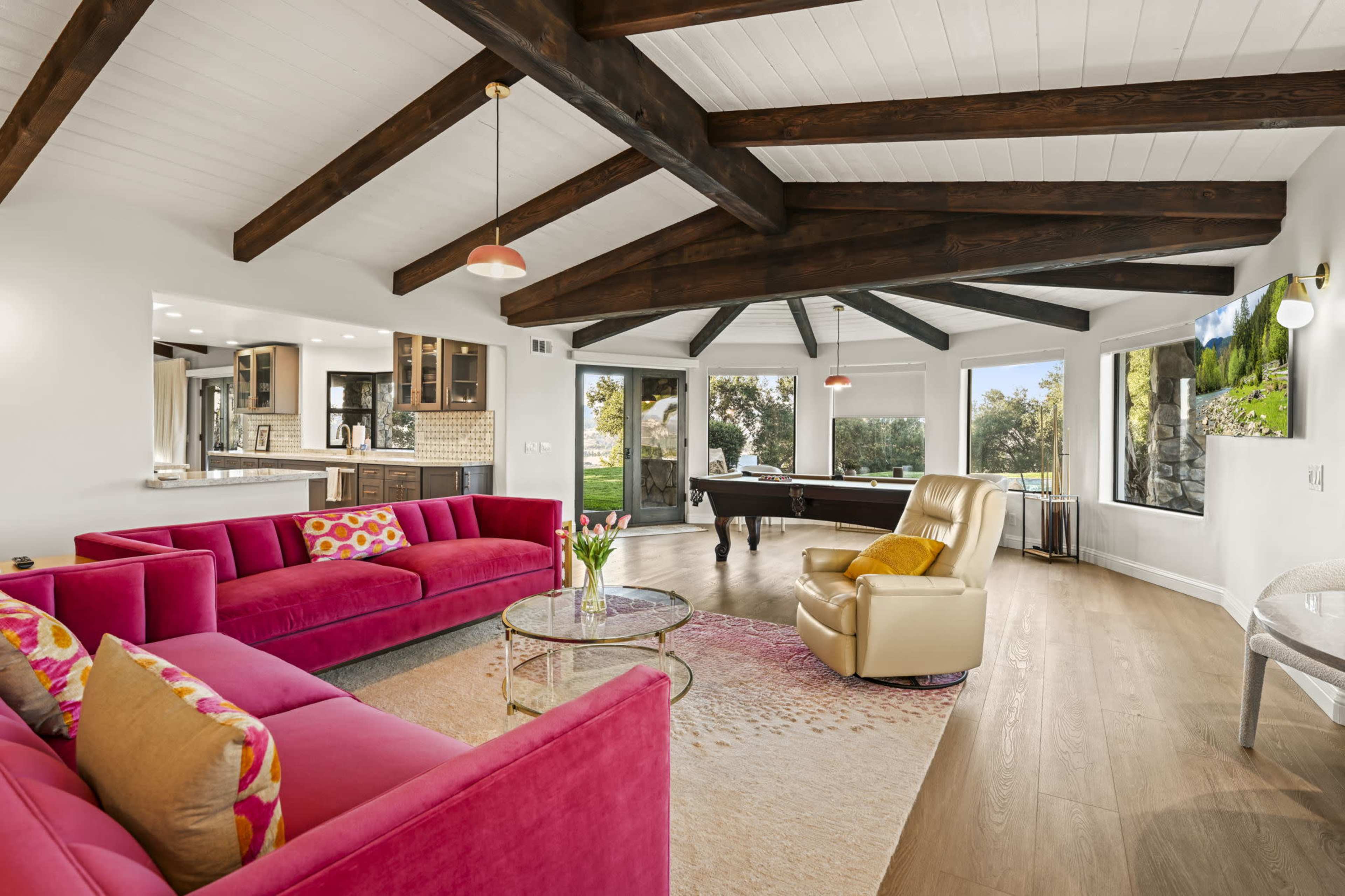 A spacious living room features a pink sectional sofa, a circular glass coffee table, and a beige recliner beneath exposed wooden beams, with large windows providing views of the outdoor landscape.