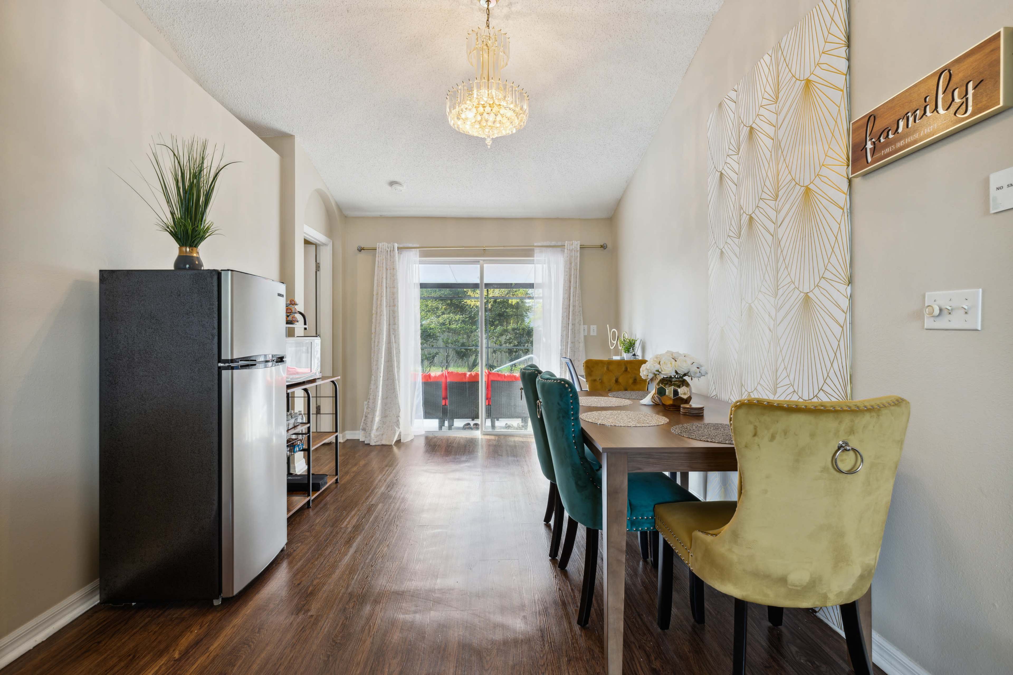 A dining area features a table with colorful chairs, a small refrigerator, and a decorative wall with a 'Family' sign, leading to a sliding glass door.