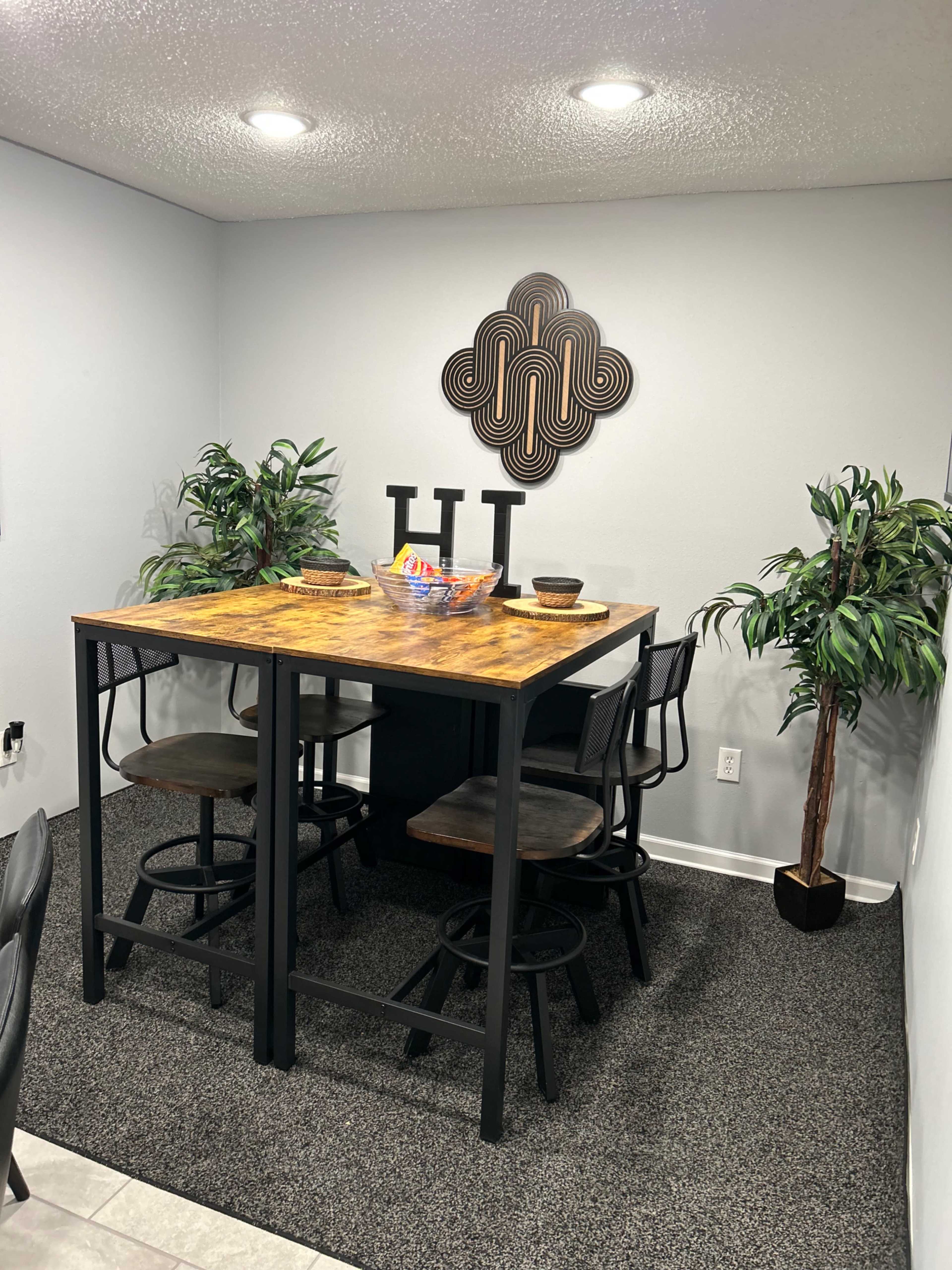 A modern dining area features a tall wooden table with four metal stools, surrounded by two decorative plants and a wall hanging design.