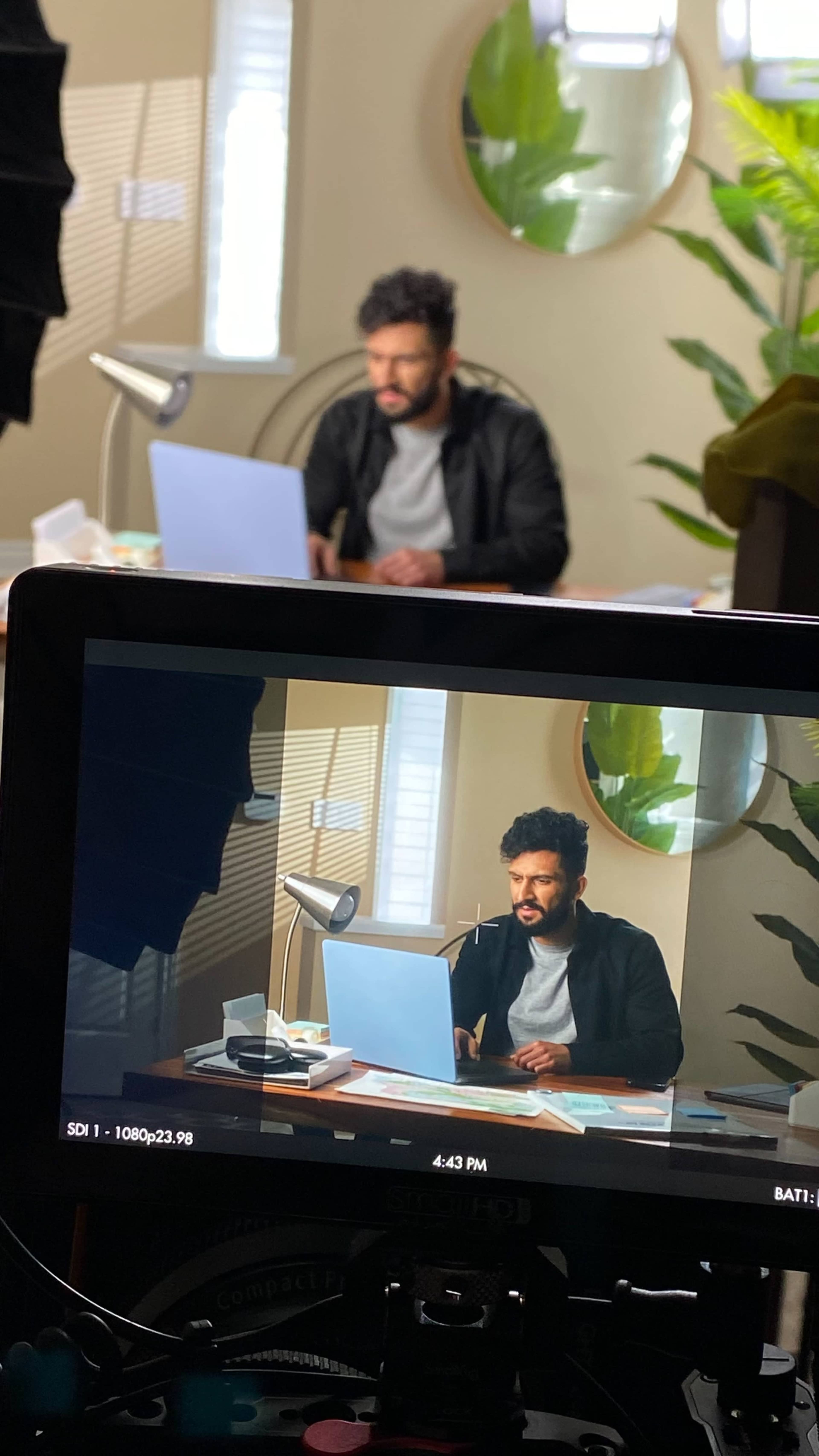 A man is sitting at a desk in front of a computer, while being filmed from a camera positioned nearby.