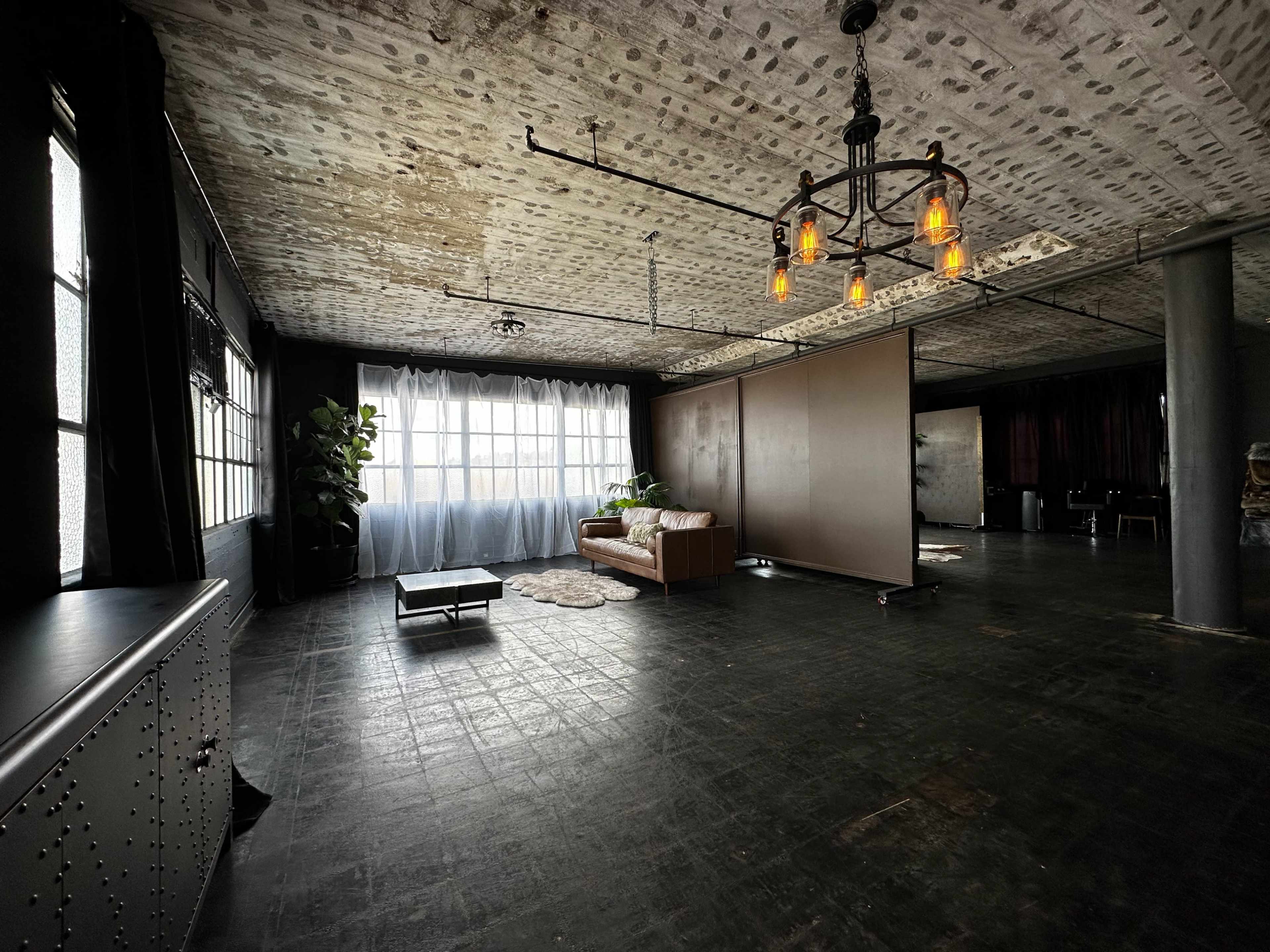 The image shows an industrial-style interior with a large window, a brown couch, a coffee table, and a minimalist design featuring concrete walls and a hanging light fixture.