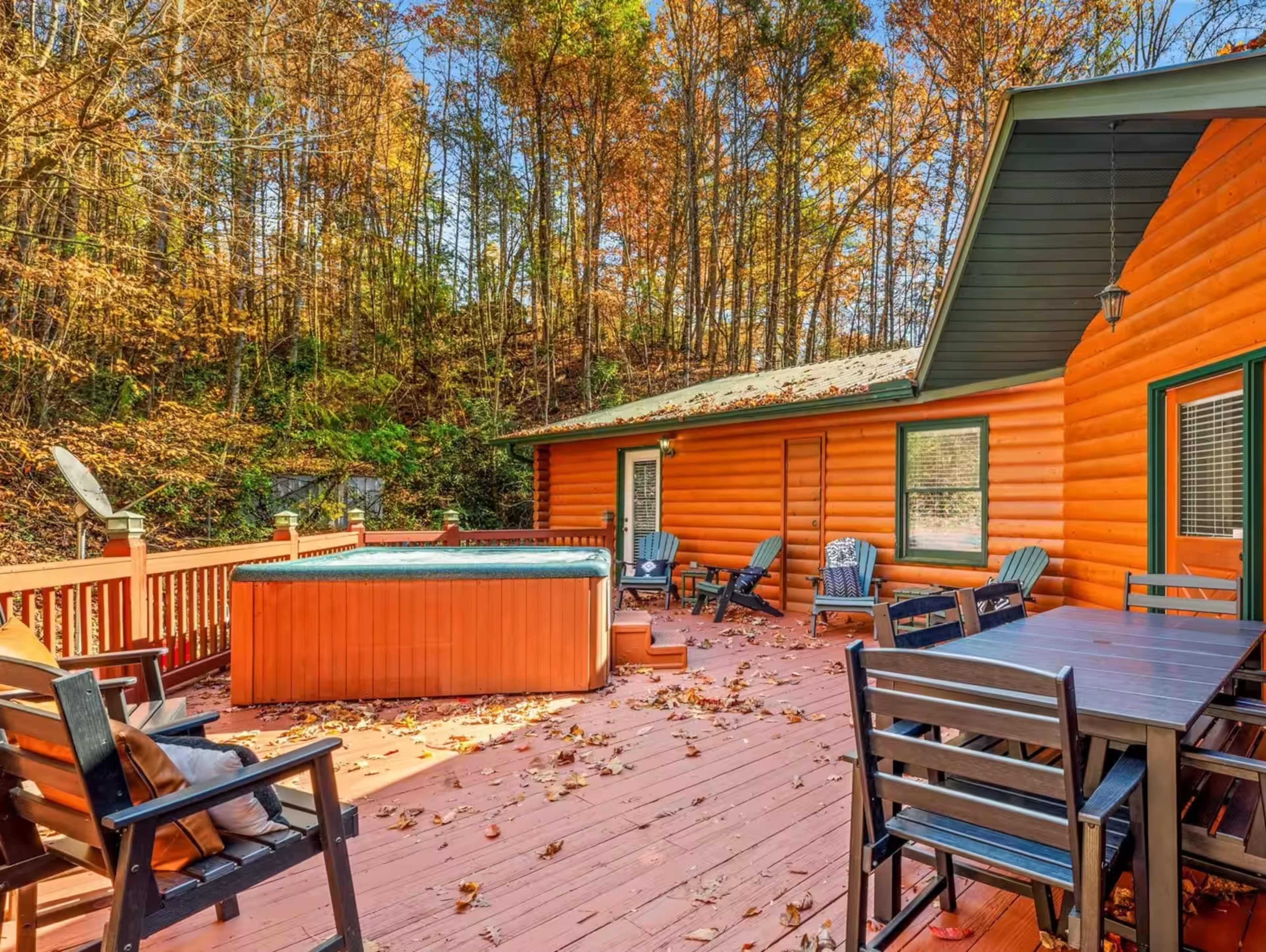 The image shows a wooden deck with seating, a hot tub, and a cabin surrounded by trees with autumn foliage.