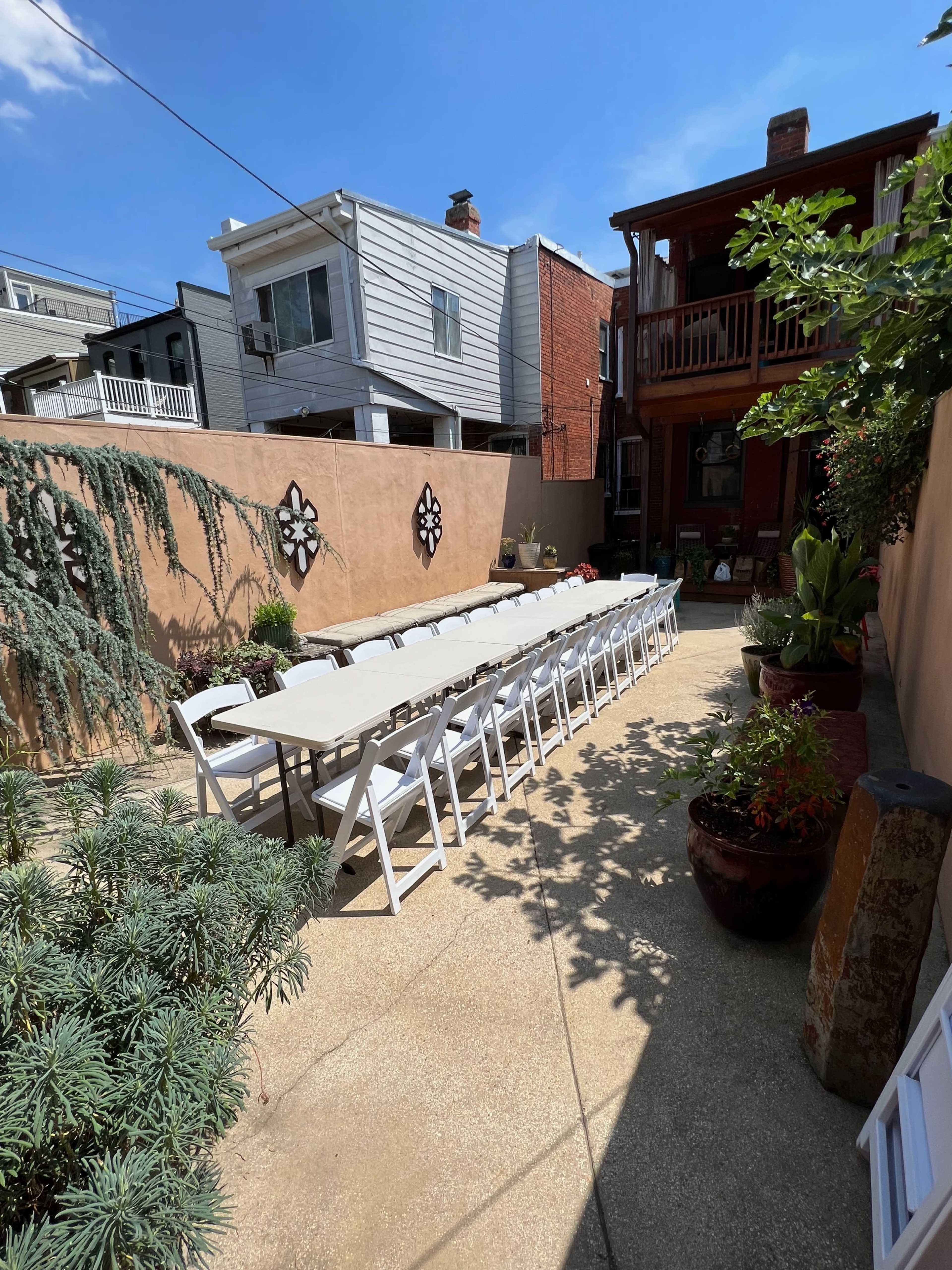 A long, white table with folding chairs is set up in a sunlit outdoor courtyard surrounded by walls and plants.
