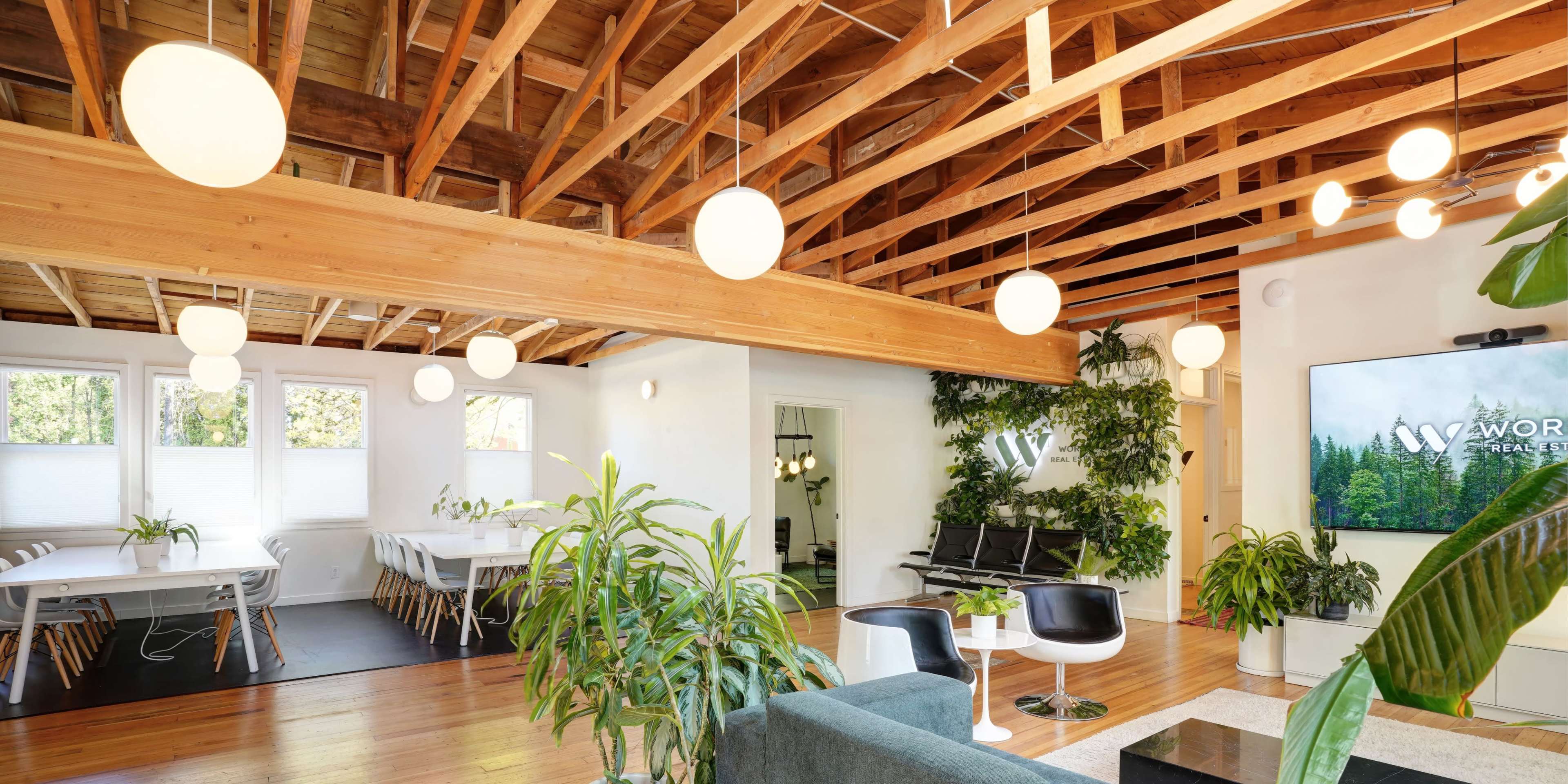 The image depicts a modern office space featuring a wooden ceiling, multiple plants, and areas for both collaboration and relaxation.