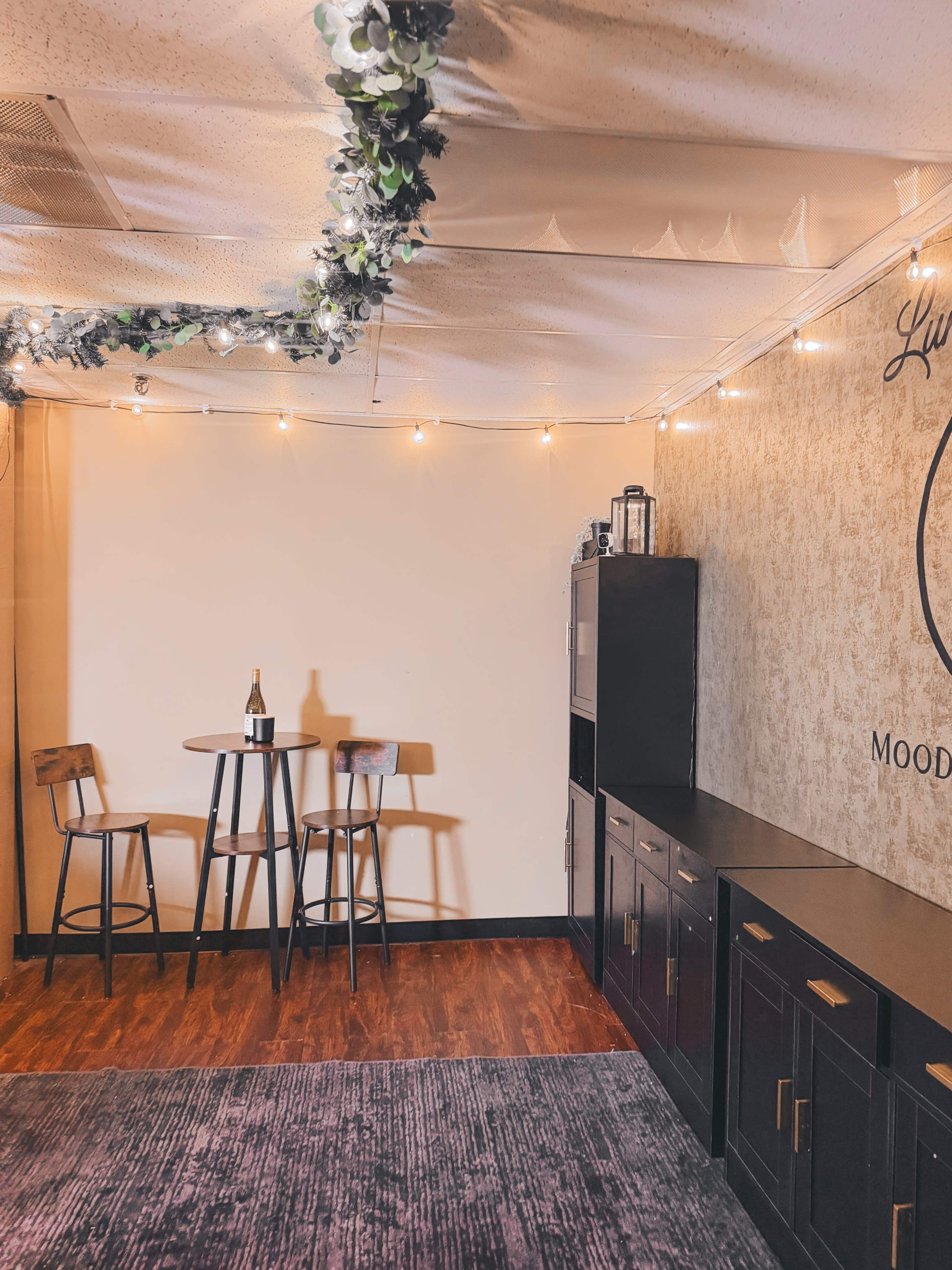 The image shows a cozy room setup with two high stools and a small table, a wine bottle on the table, and lighting fixtures along the ceiling.