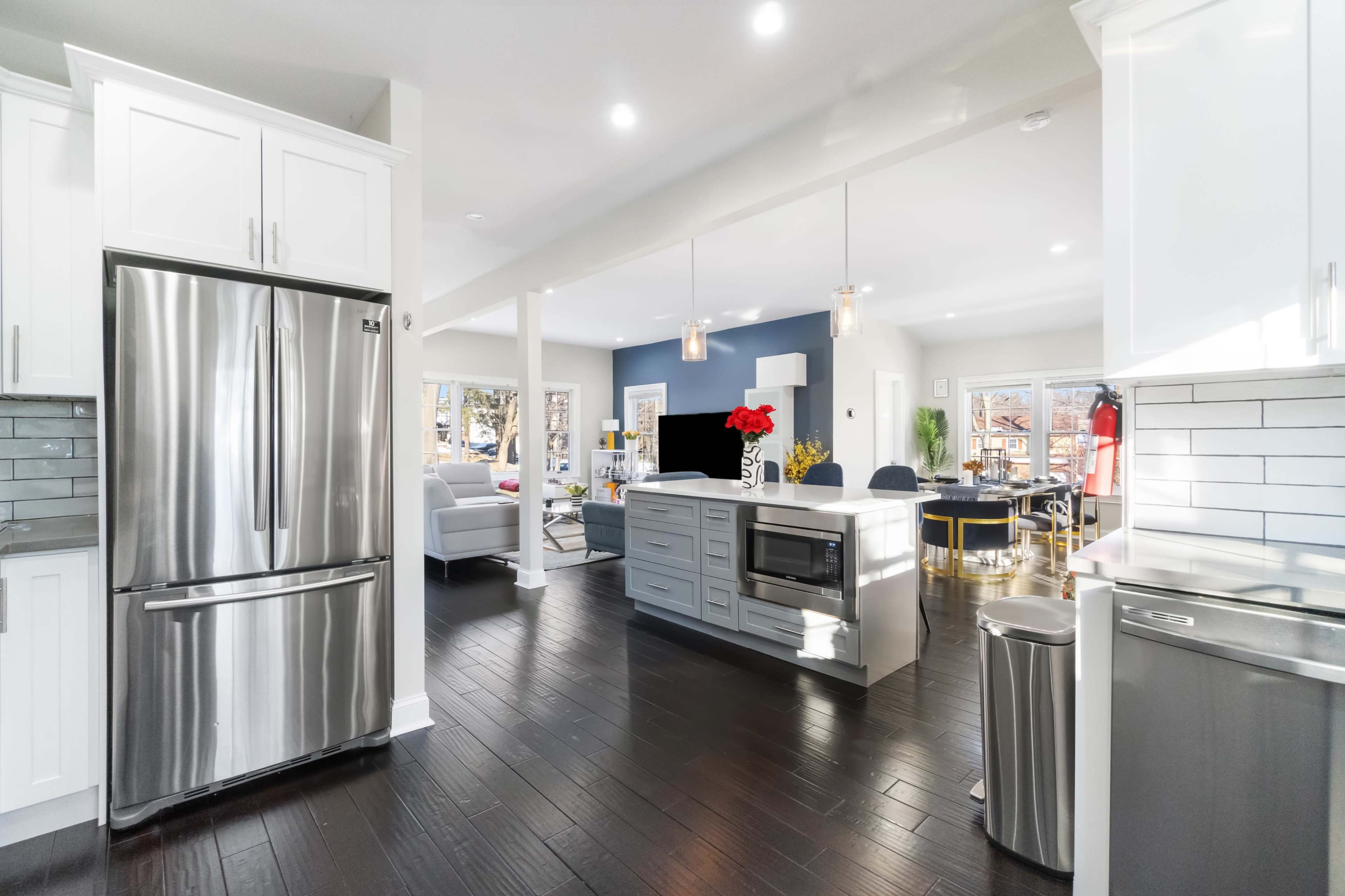 The image shows a modern kitchen with stainless steel appliances, an island with a microwave and seating area, and a view into a bright living space with a blue accent wall.