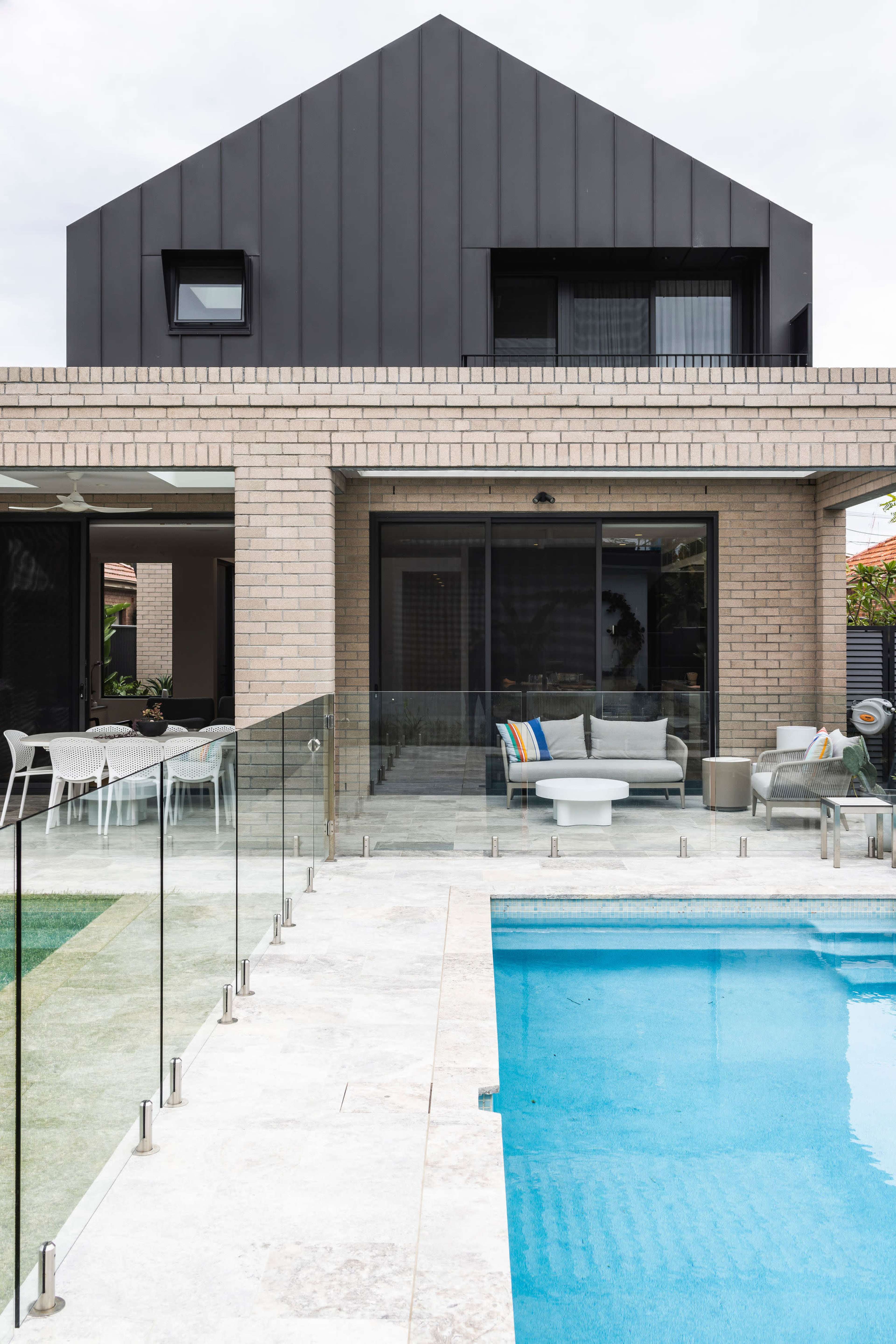 A modern house with a dark, sloped roof and large glass doors overlooks a swimming pool surrounded by a glass railing.