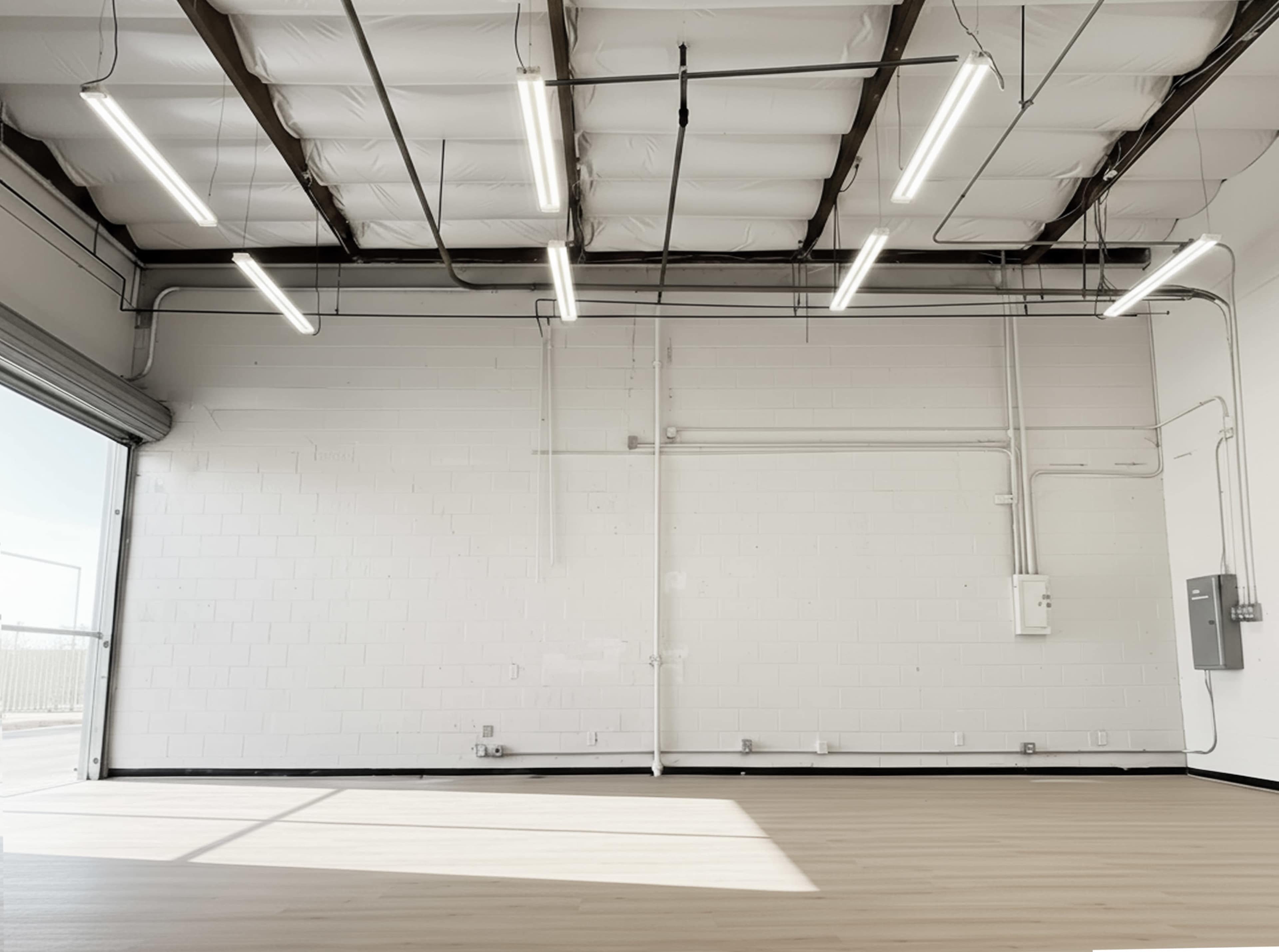 The image shows a spacious, empty room with a white wall, wooden flooring, and industrial ceiling lights.
