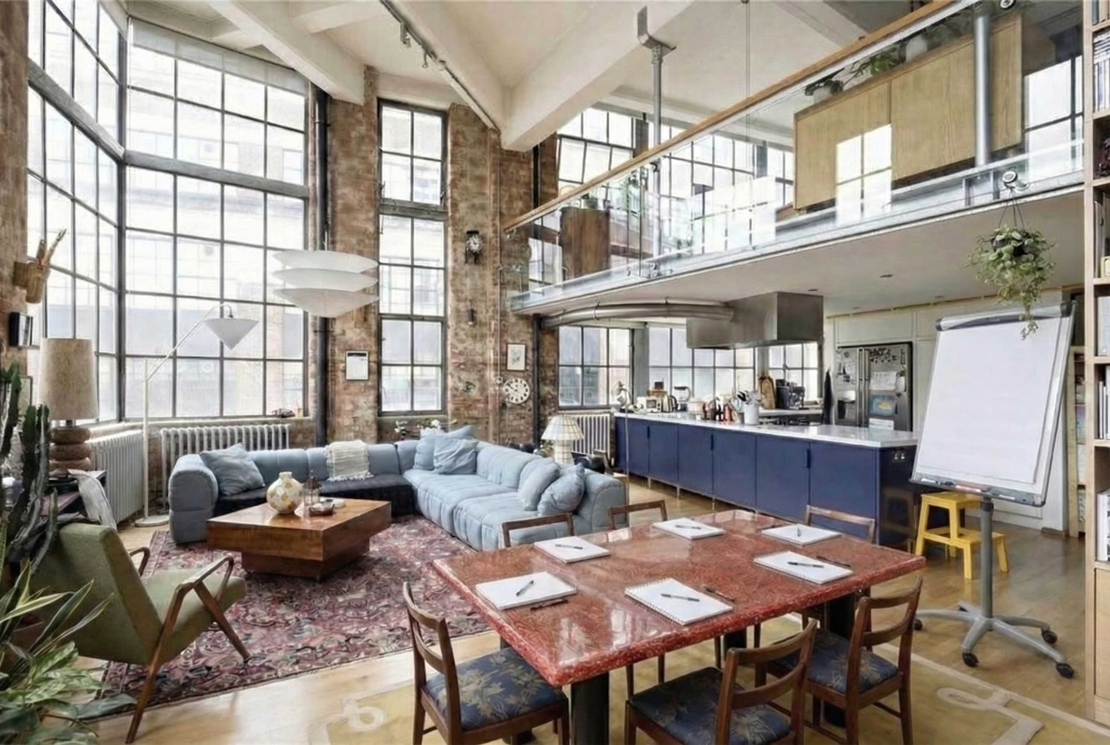 The image shows a spacious, airy loft-style living area featuring large windows, a comfortable seating area, and a dining table surrounded by chairs.