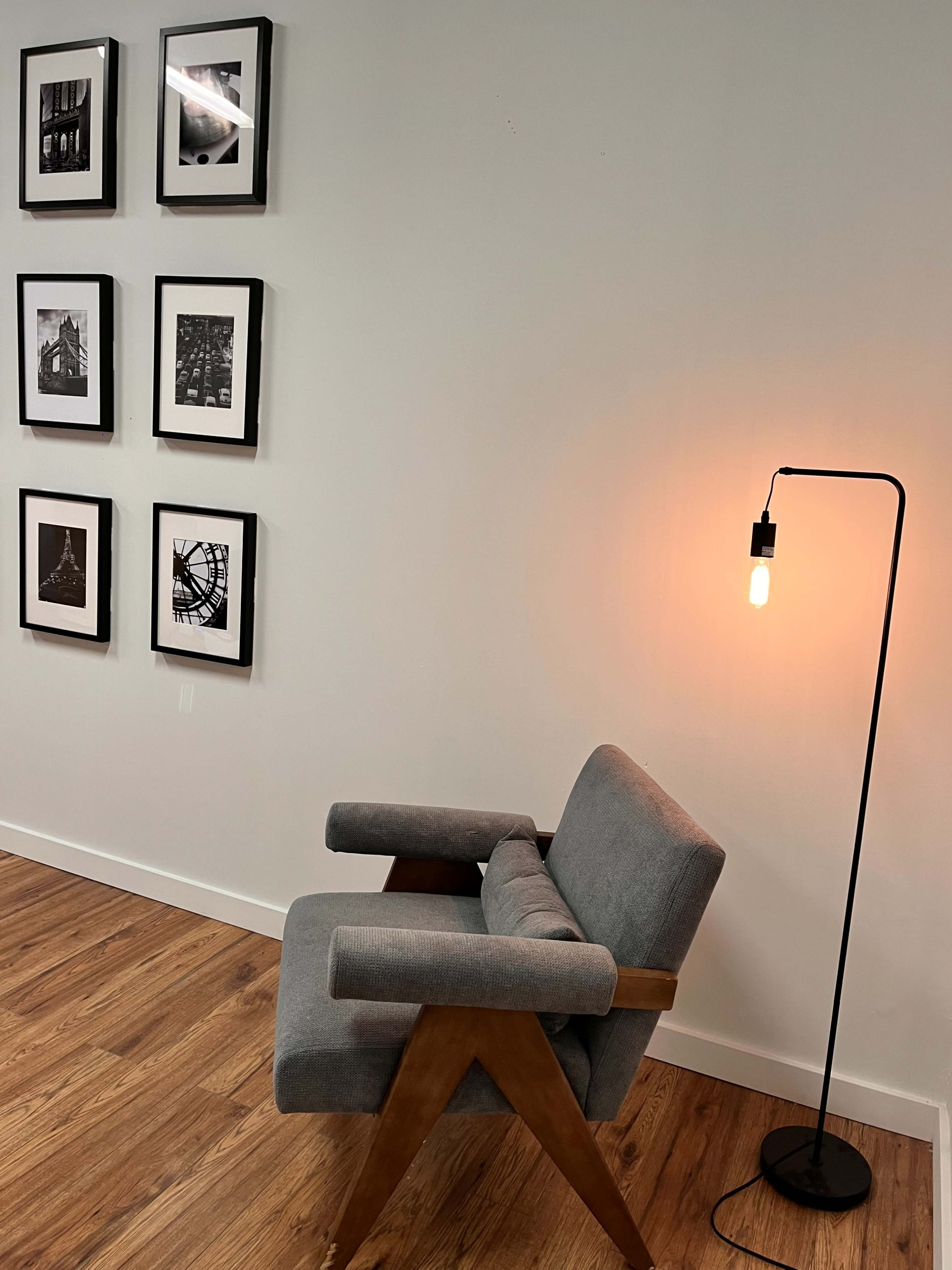 A gray armchair is positioned beside a floor lamp, in front of a wall adorned with framed black-and-white photographs.