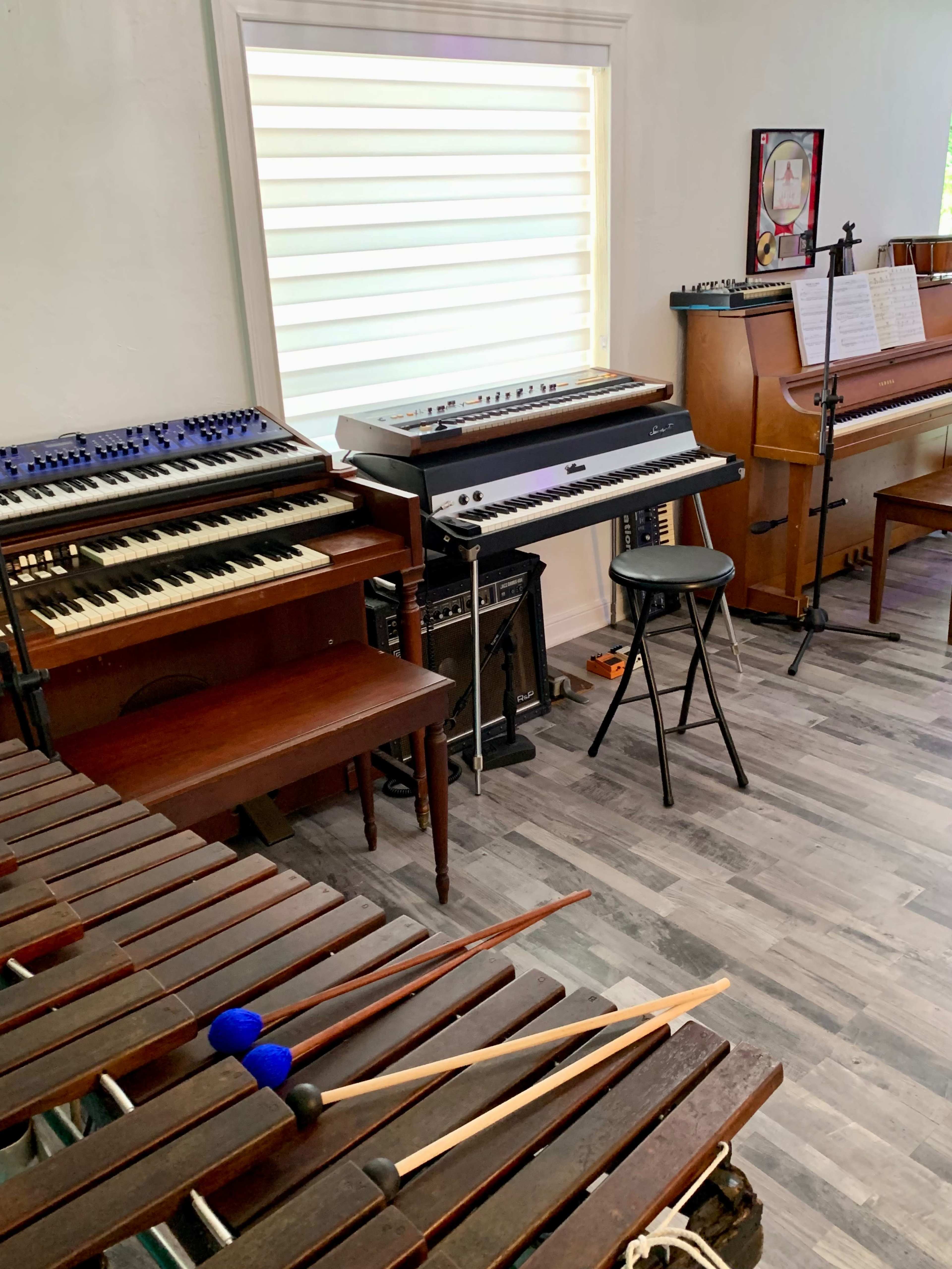 The image shows a music room with a xylophone in the foreground, various keyboards, a piano, and sheet music arranged on a stand.