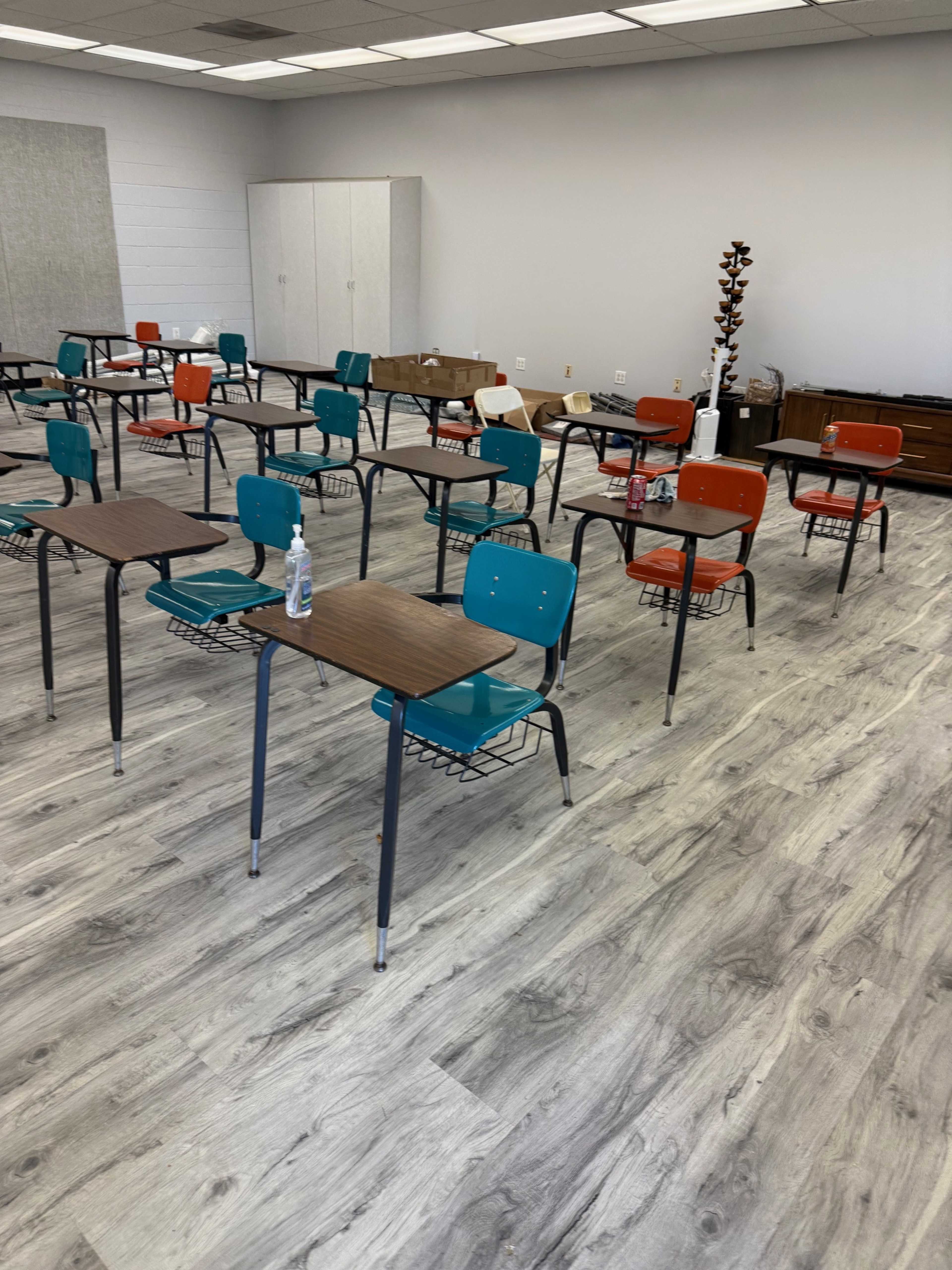 A classroom with several desks and chairs arranged in rows, featuring a mix of orange and teal seating, and a wood cabinet in the background.